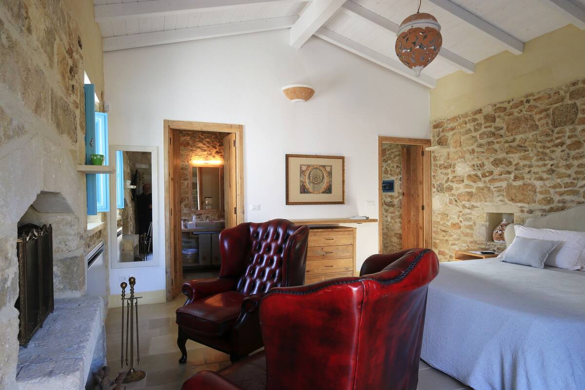 Un dormitorio con pared de piedra, una cama, un sillón de cuero rojo, un aparador de madera y un espejo.