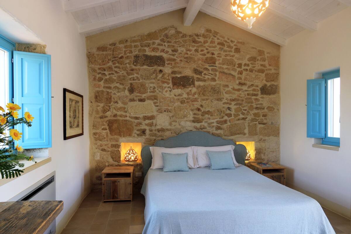 Un dormitorio con pared de piedra, una cama con cabecera azul, dos mesitas de noche con lámparas y una ventana con contraventanas azules.