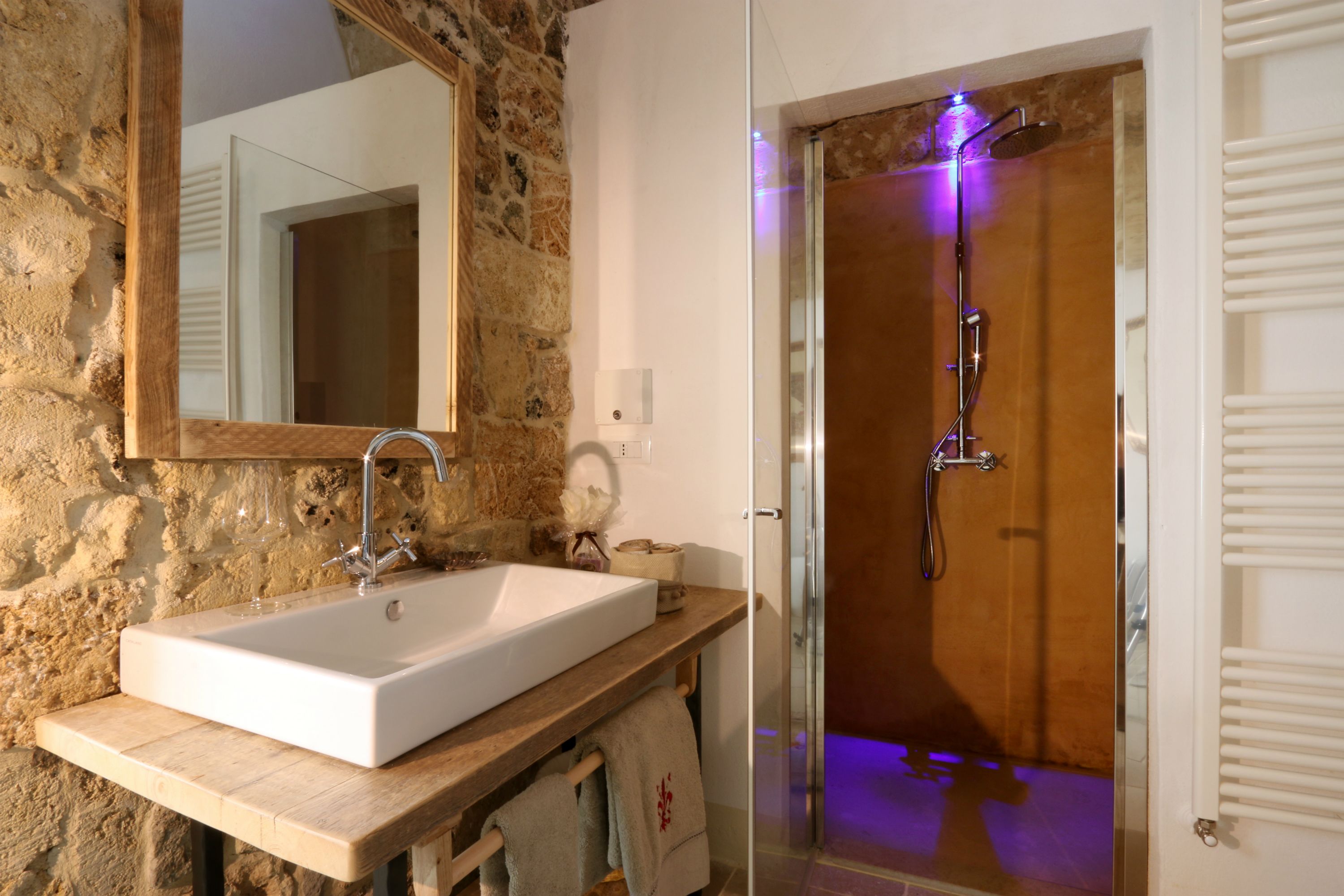 A bathroom with a stone wall, a wooden vanity, a large mirror, a white sink, a shower with a glass door, and a towel rack.