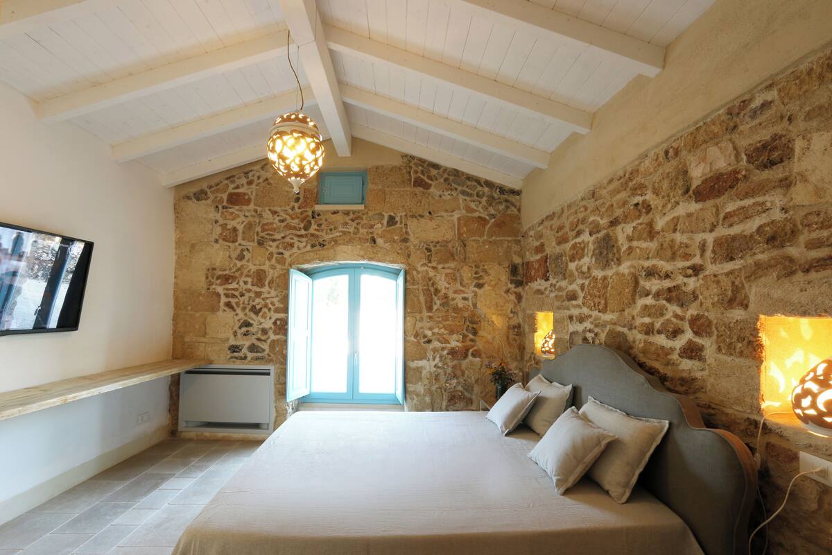 Un dormitorio con pared de piedra, una cama con almohadas, una lámpara colgante y una ventana con vistas.