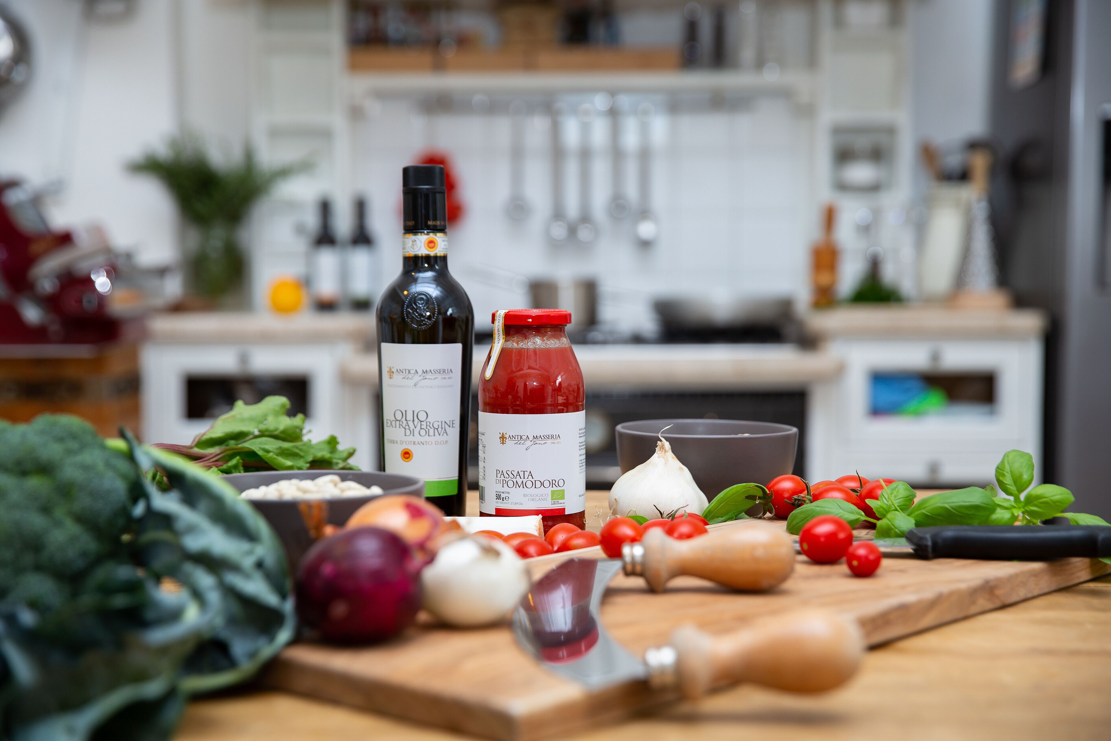 Una encimera de cocina con varias verduras, una botella de aceite de oliva, una botella de passata de tomate y una tabla de cortar.