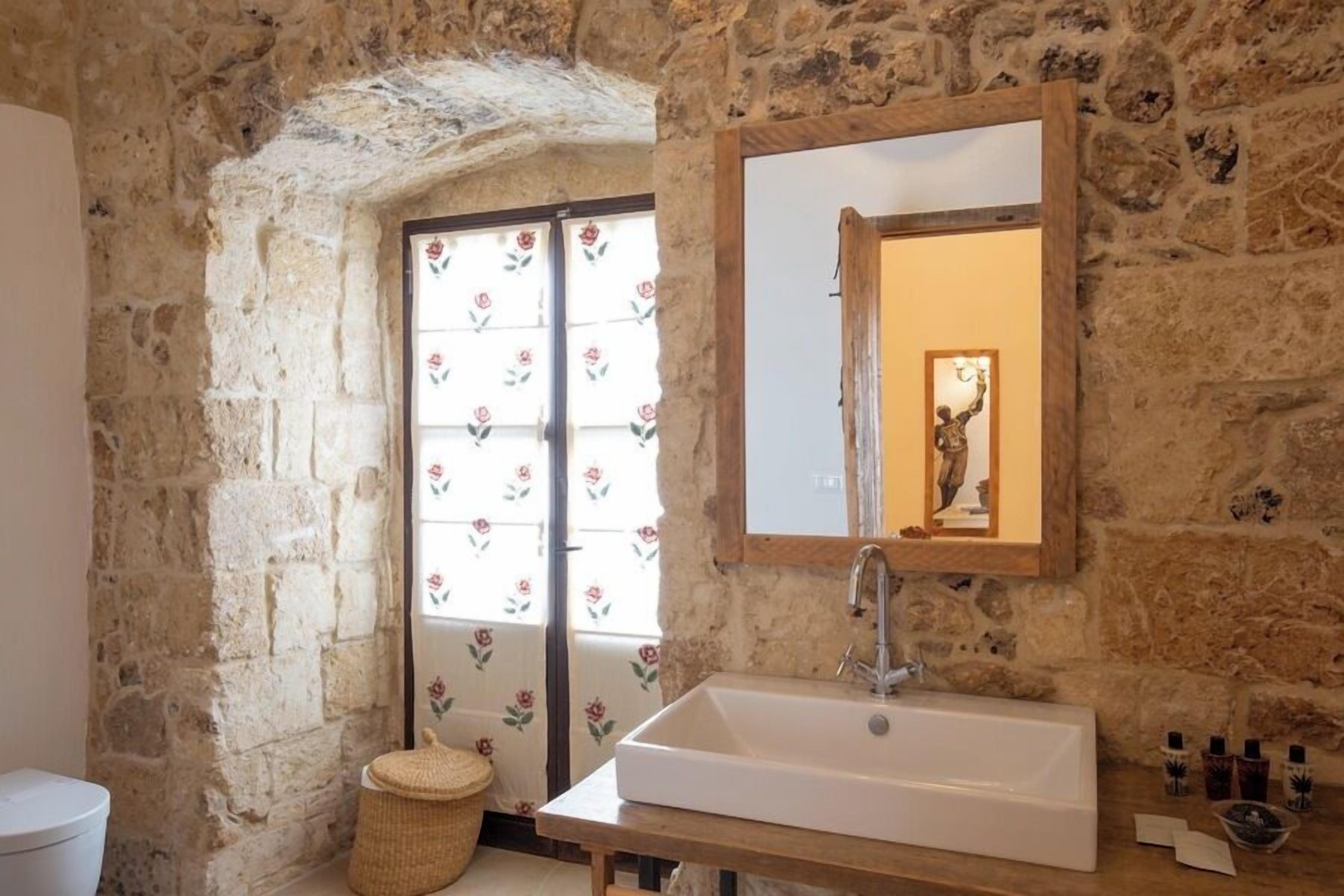 A bathroom with a stone wall, a white sink, a mirror, and a window with floral curtains.