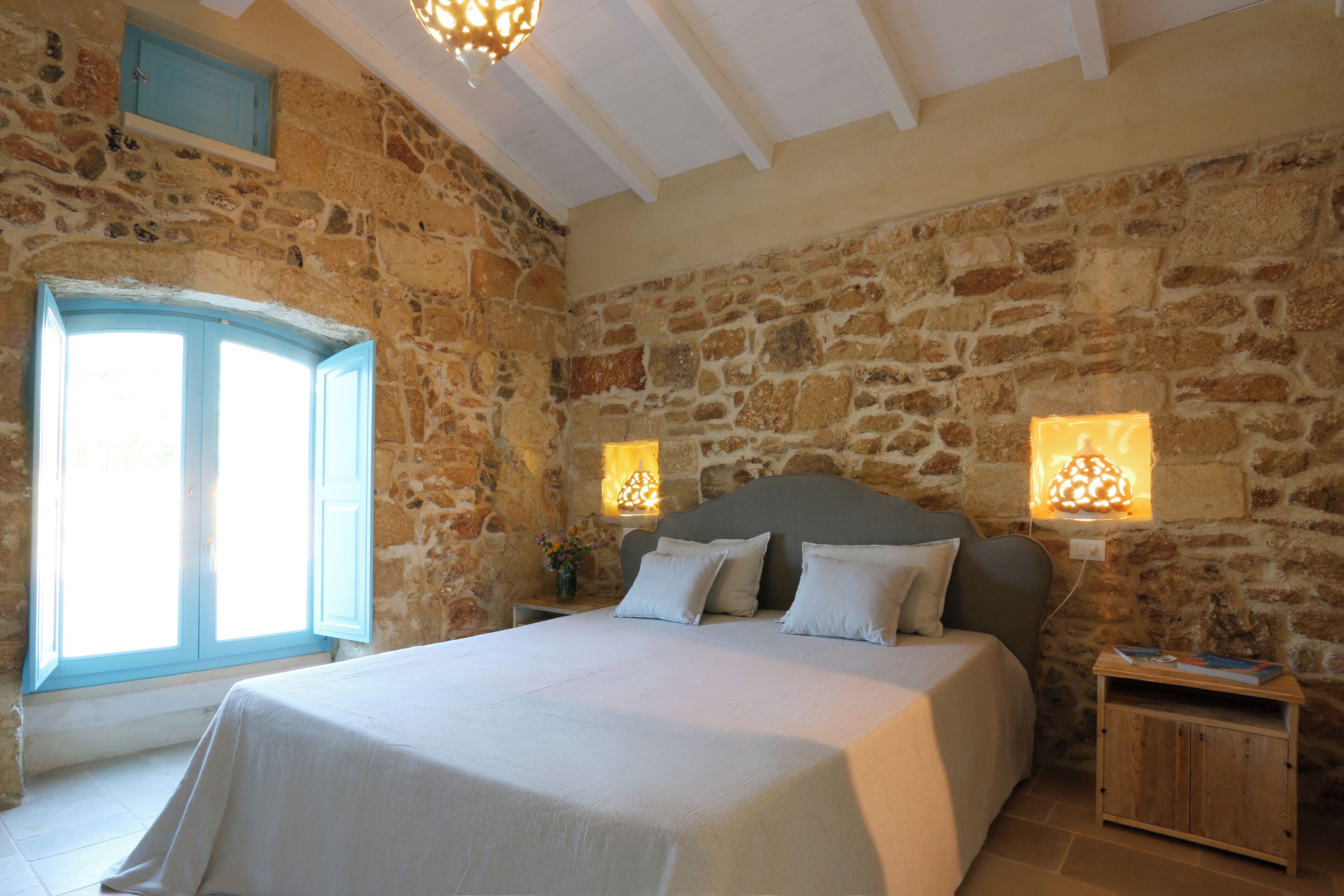 Un dormitorio con pared de piedra, una cama con ropa de cama blanca y dos lámparas de pared.