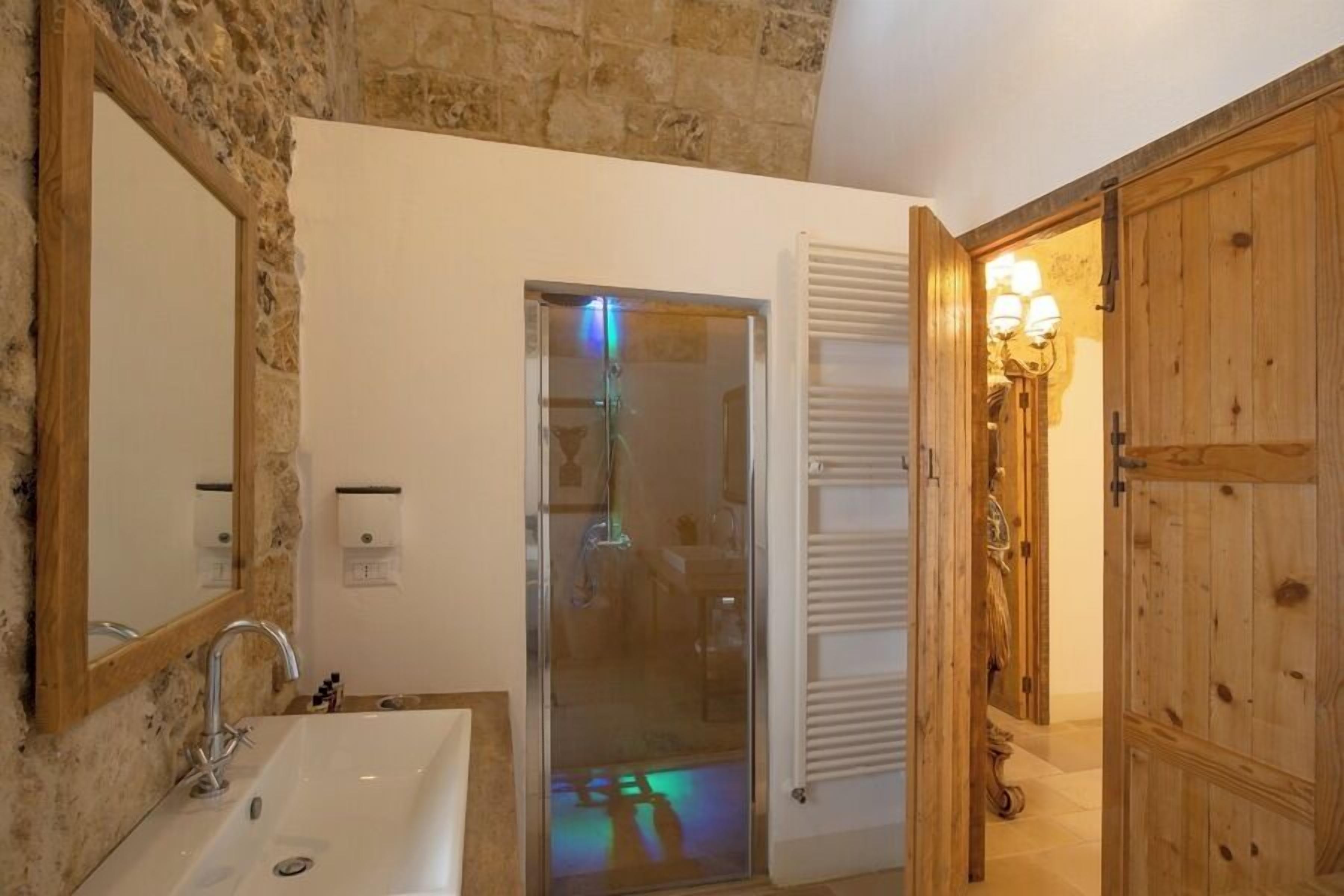 Une salle de bain avec un mur en pierre, un lavabo blanc, une cabine de douche en verre et des portes en bois.