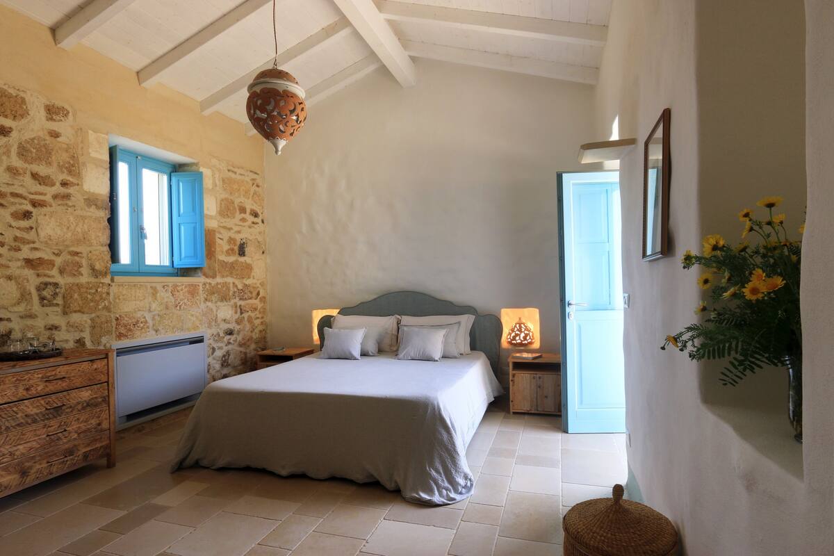 Un dormitorio con pared de piedra, cama con ropa blanca, cómoda de madera y lámpara colgante.