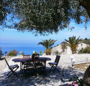 A stone patio with a round table and four chairs, overlooking a coastal landscape with the sea and hills in the background. - La Casa D' Irene (Lefkada)