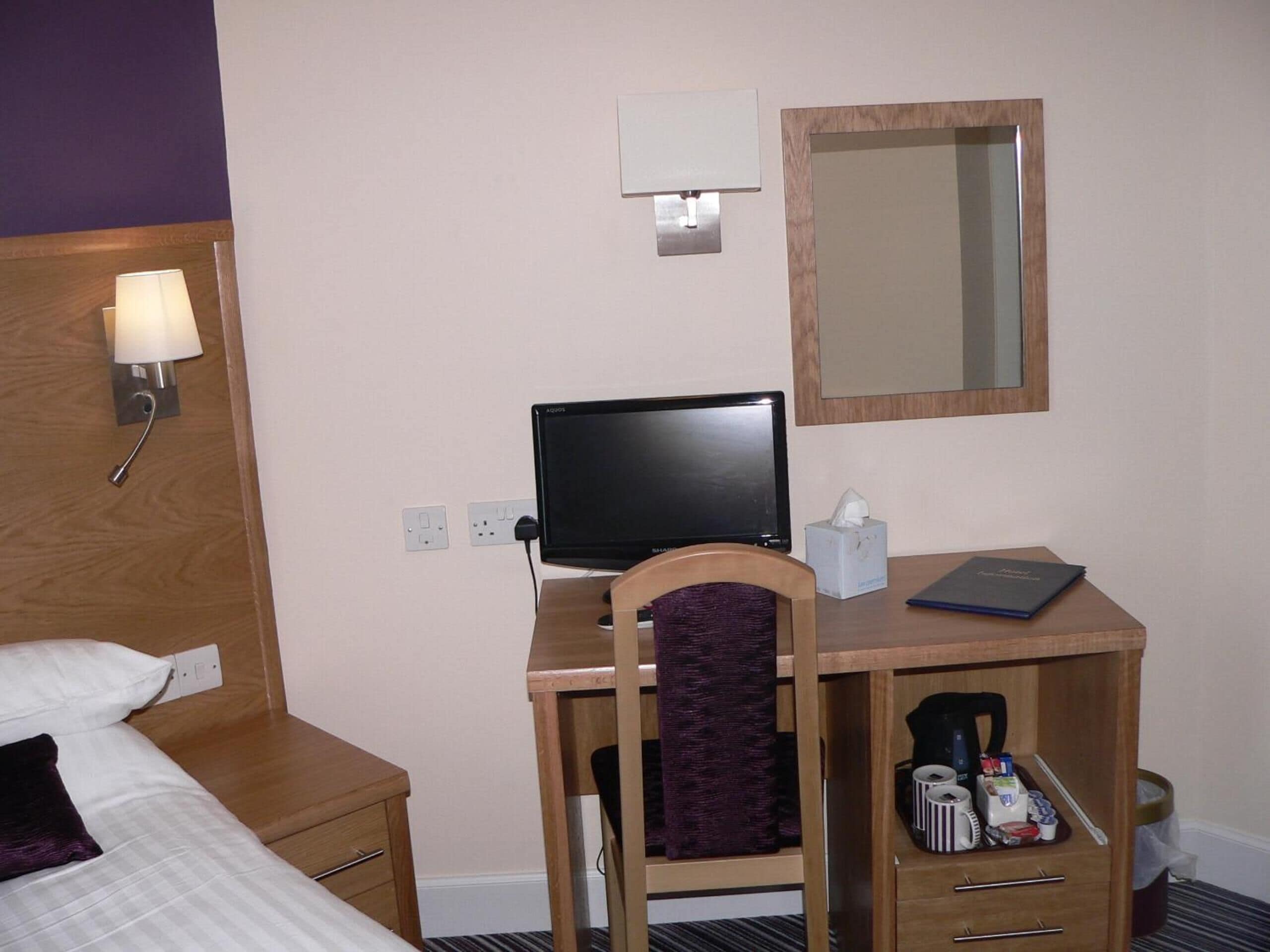 A hotel room with a bed, a desk with a computer, a mirror, and a bedside lamp.