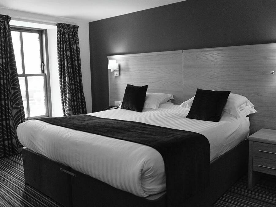 A hotel room with a large bed, two bedside tables, a nightstand, a window with curtains, and a headboard with wall-mounted lights.