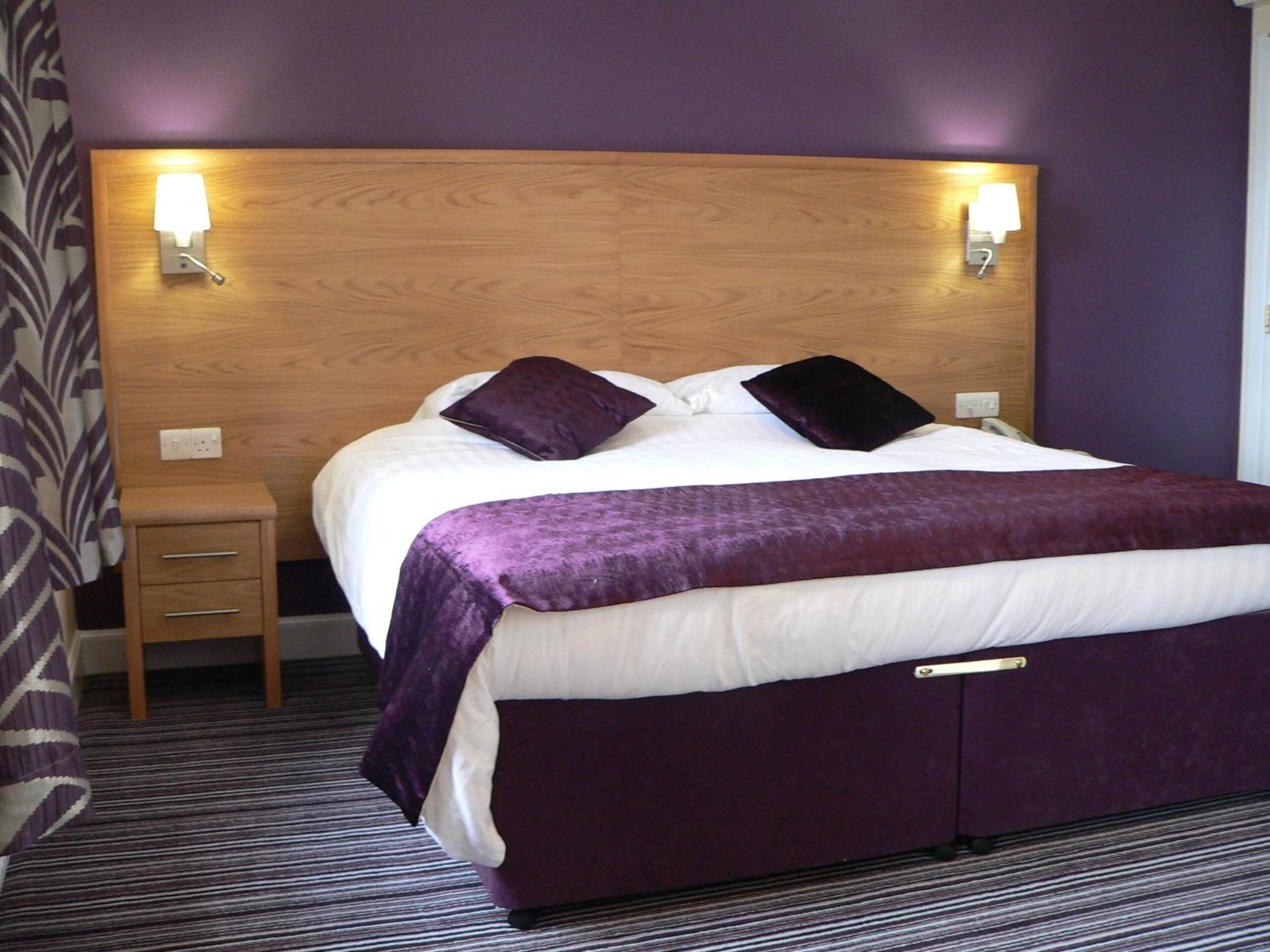 Una habitación de hotel con cabecera de madera, dos lámparas de noche, una cama con ropa de cama blanca y morada, una mesita de noche pequeña y una alfombra a rayas.