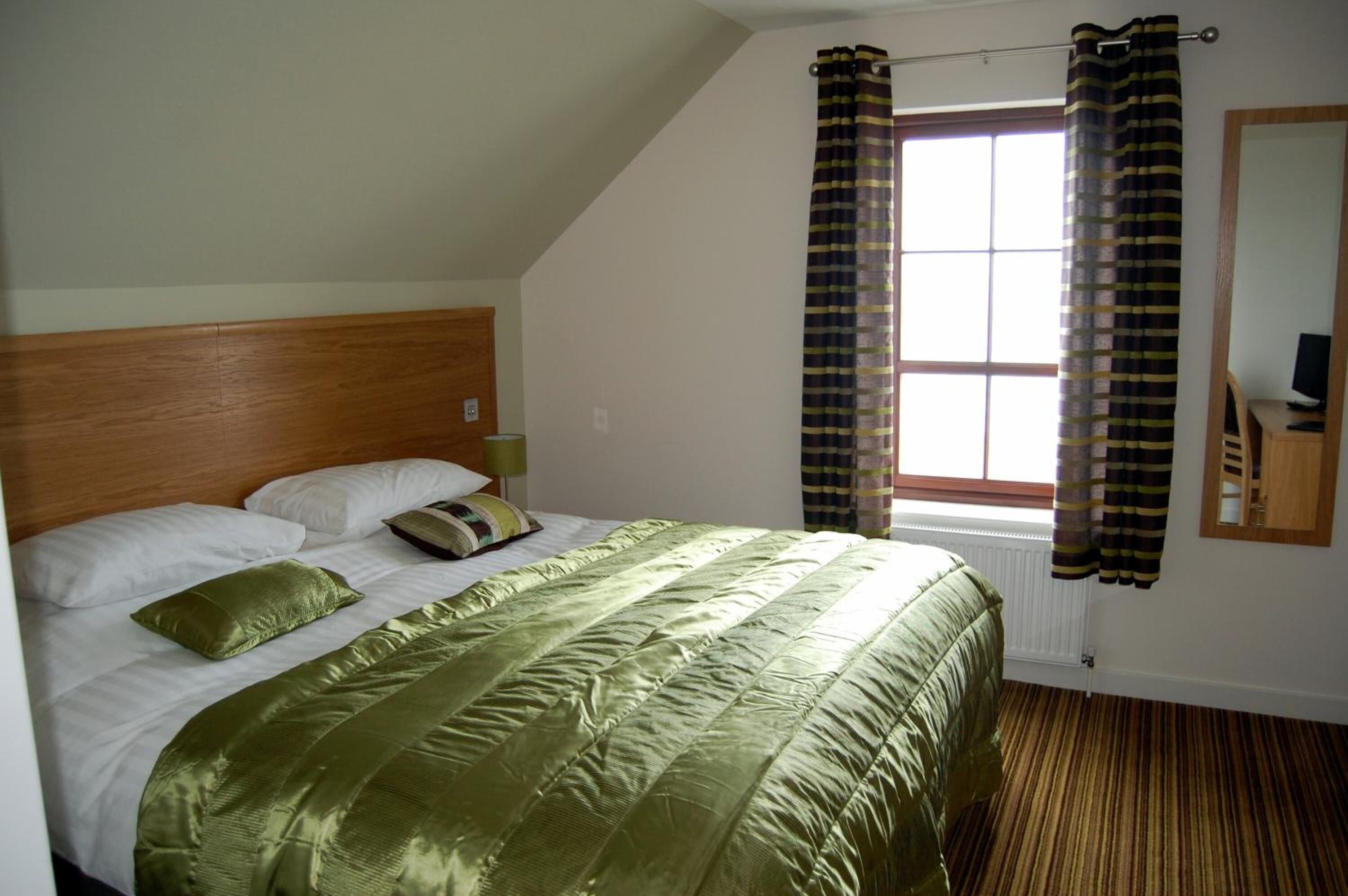 A bedroom with a bed, a wooden headboard, a window with curtains, and a mirror on the wall.
