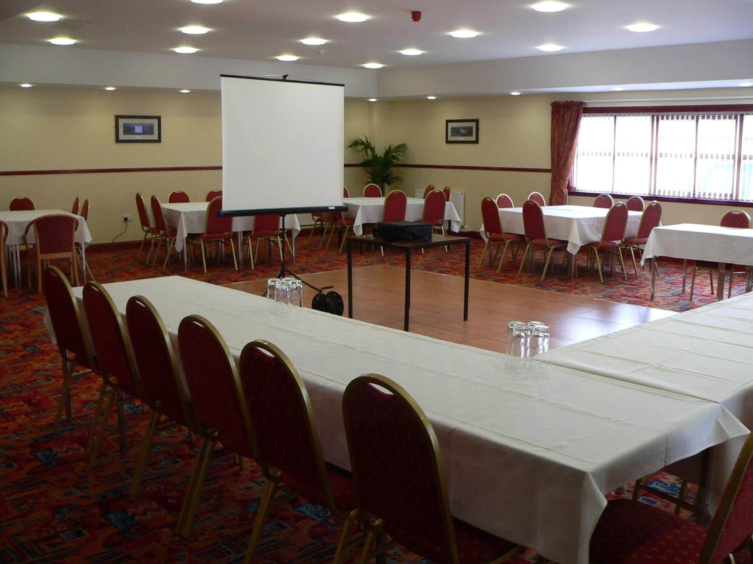 A conference room with rows of tables and chairs, a projector screen, and a carpeted floor.