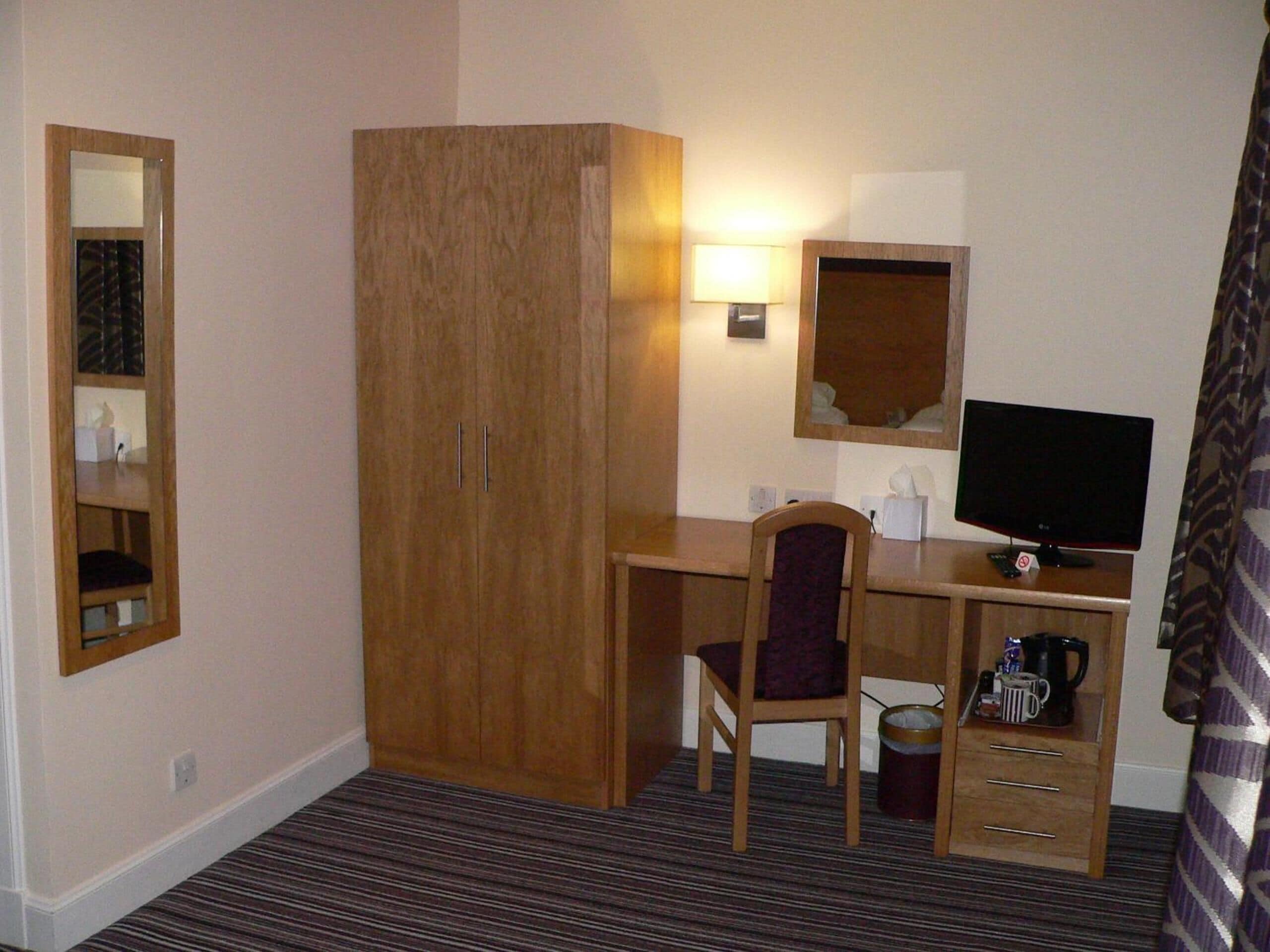 Habitación de hotel con armario de madera, un escritorio con silla, televisor y un pequeño cubo de basura.