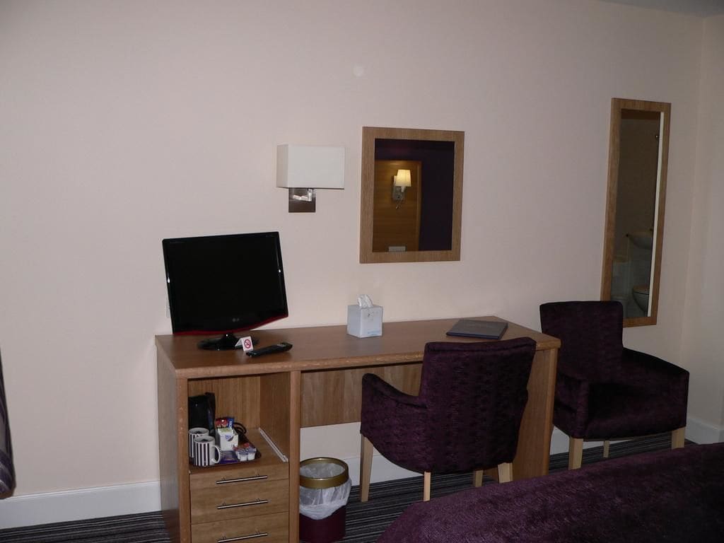 Habitación de hotel con un escritorio de madera, una computadora, dos sillas moradas, un espejo y un cesto de basura.