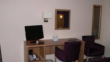 A hotel room with a wooden desk, a computer, two purple chairs, a mirror, and a trash bin.