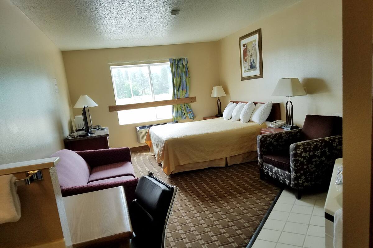 A hotel room with a bed, two chairs, a desk, a lamp, a painting, and a window.