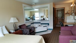 A hotel room with a large bed, a desk with a lamp, a seating area with a chair, a small table, and a bathroom with a sink and mirror.