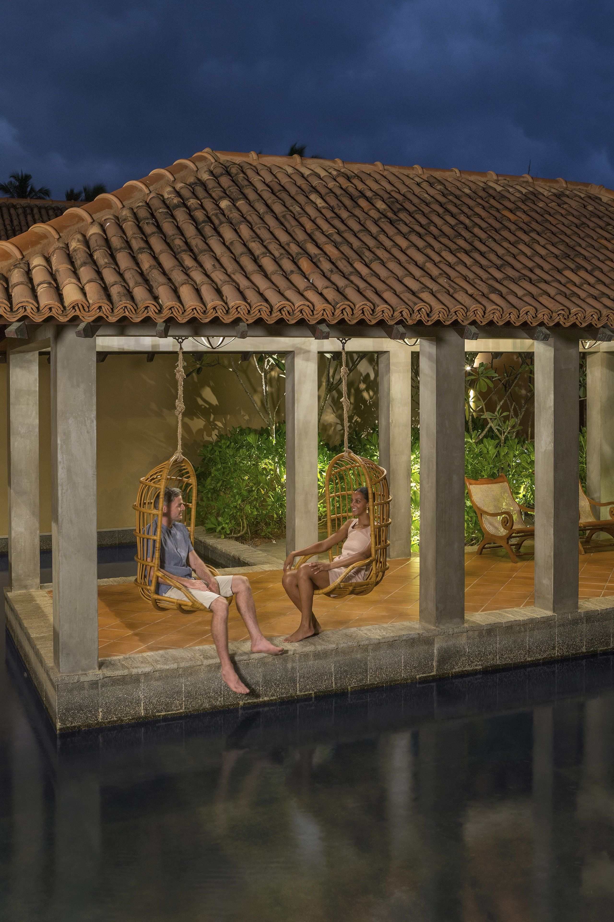 A covered outdoor seating area with two wicker swing chairs and a lounge chair, overlooking a pool.