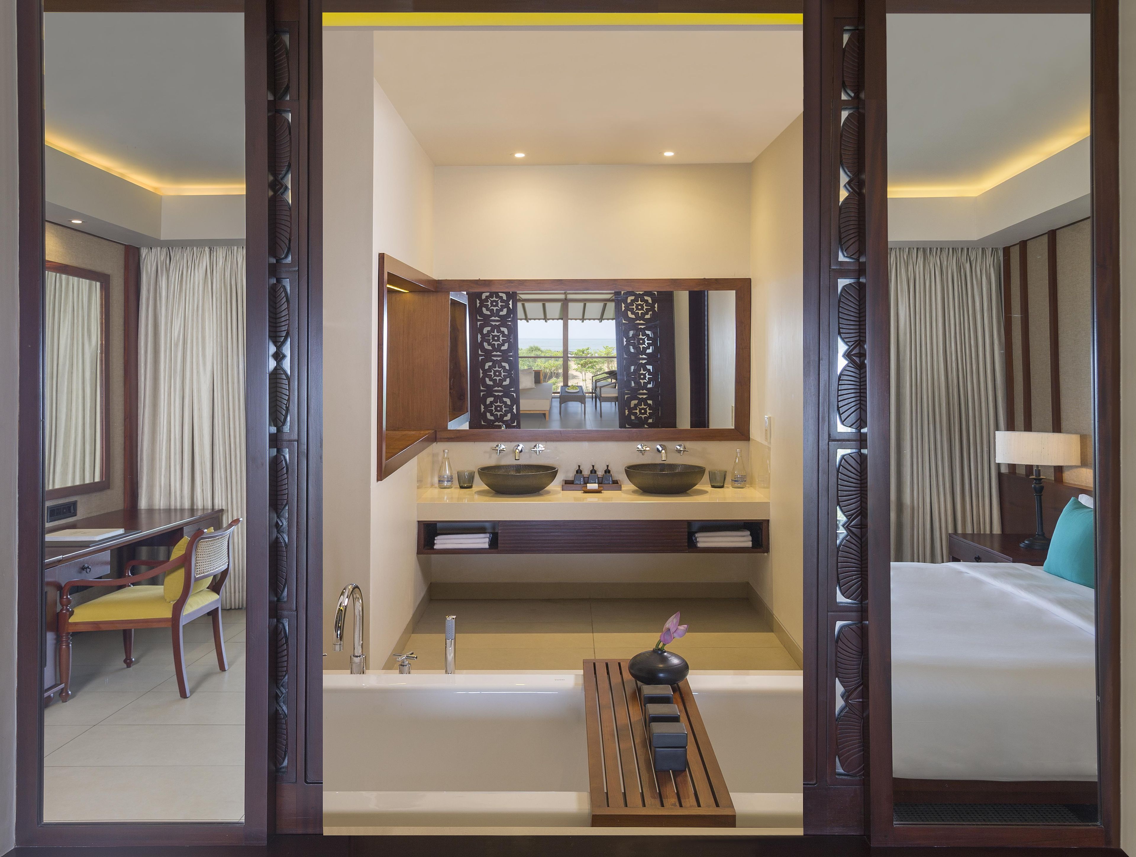 A hotel room with a large mirror, a bathtub, a vanity with a bowl sink, a desk with a chair, and a bed.