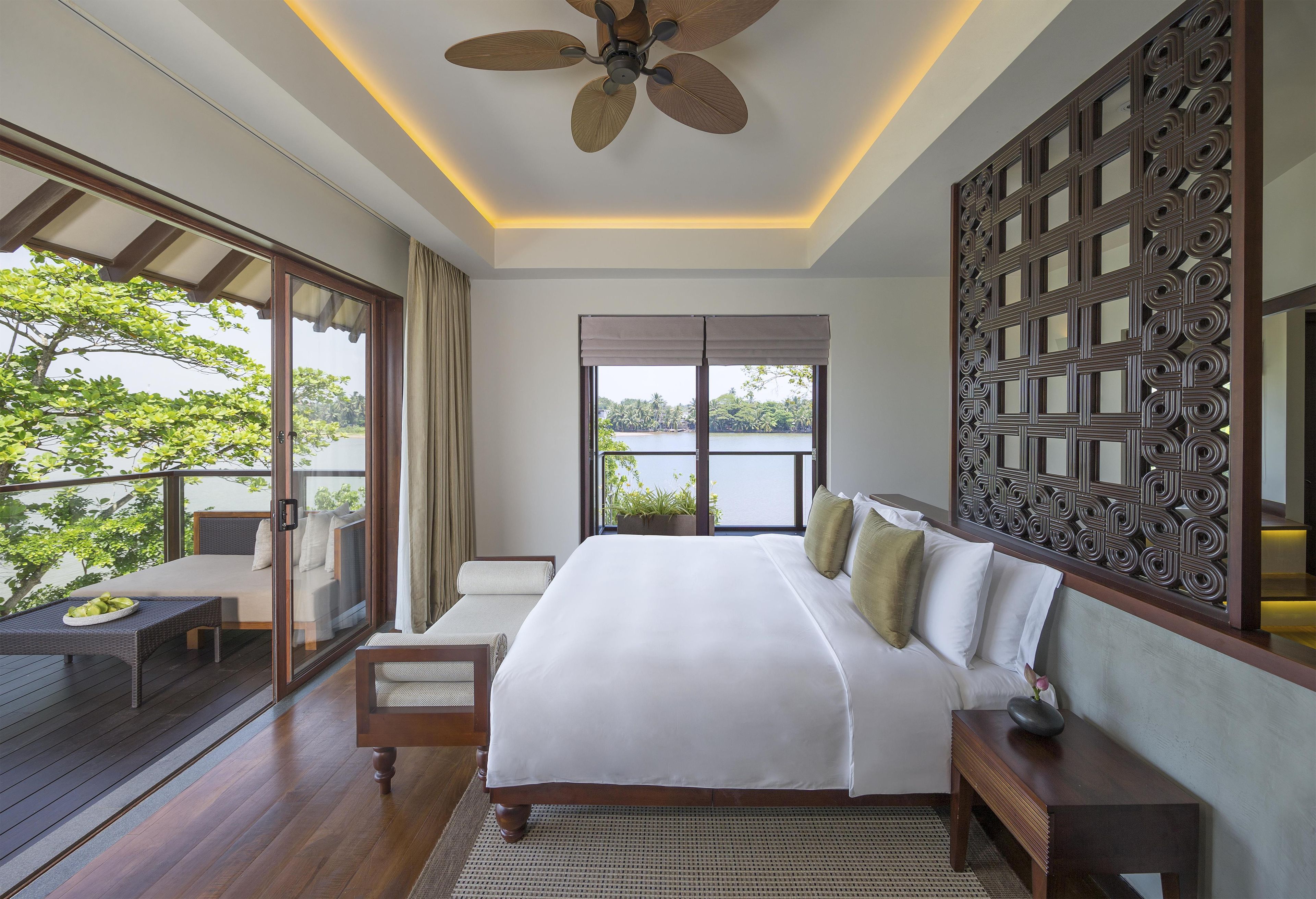 Un dormitorio con una cama grande, ventilador de techo, vista a un lago y un balcón con mobiliario exterior.
