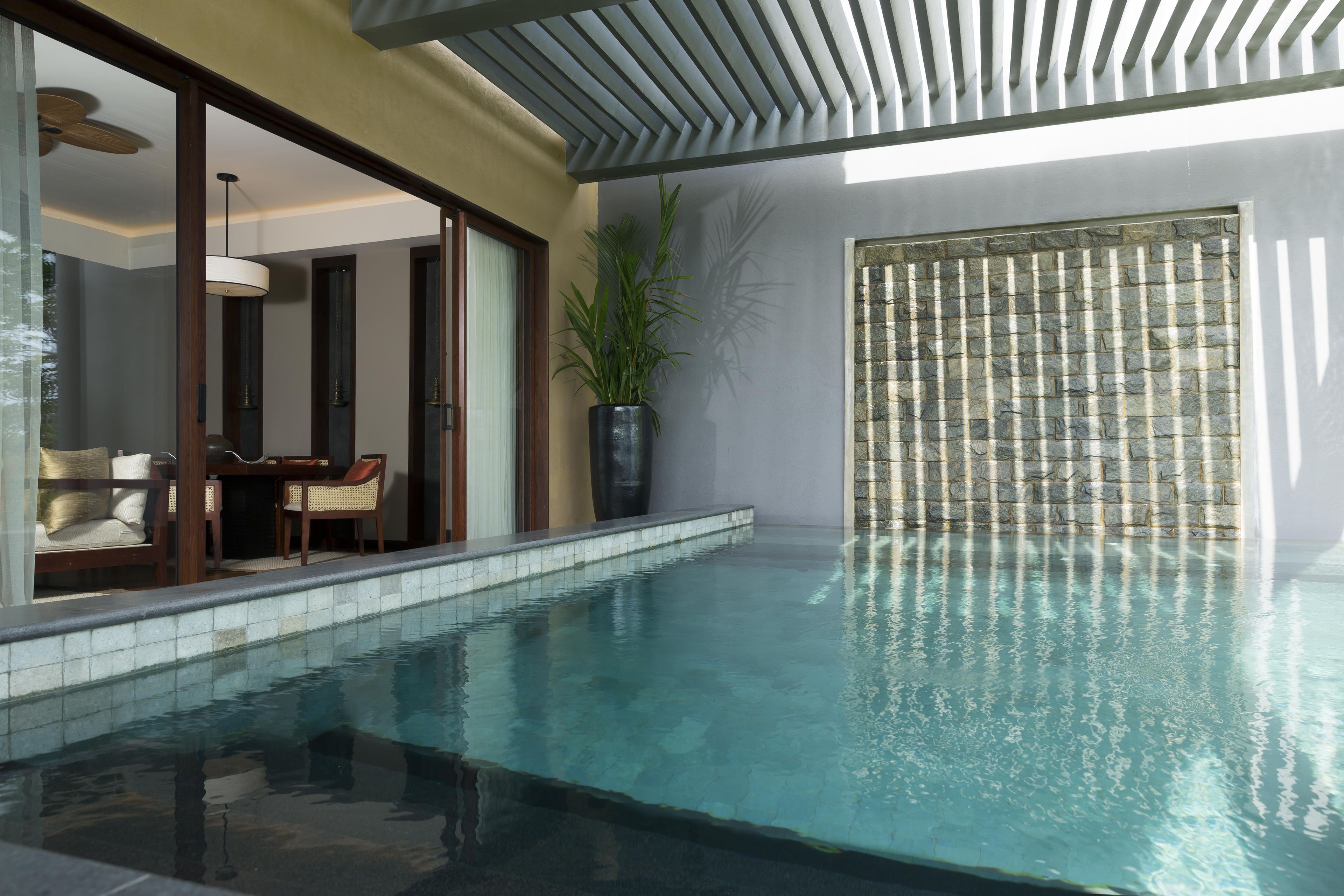 Una piscina interior moderna con agua cristalina, una zona de estar con sillones y una planta grande en maceta.