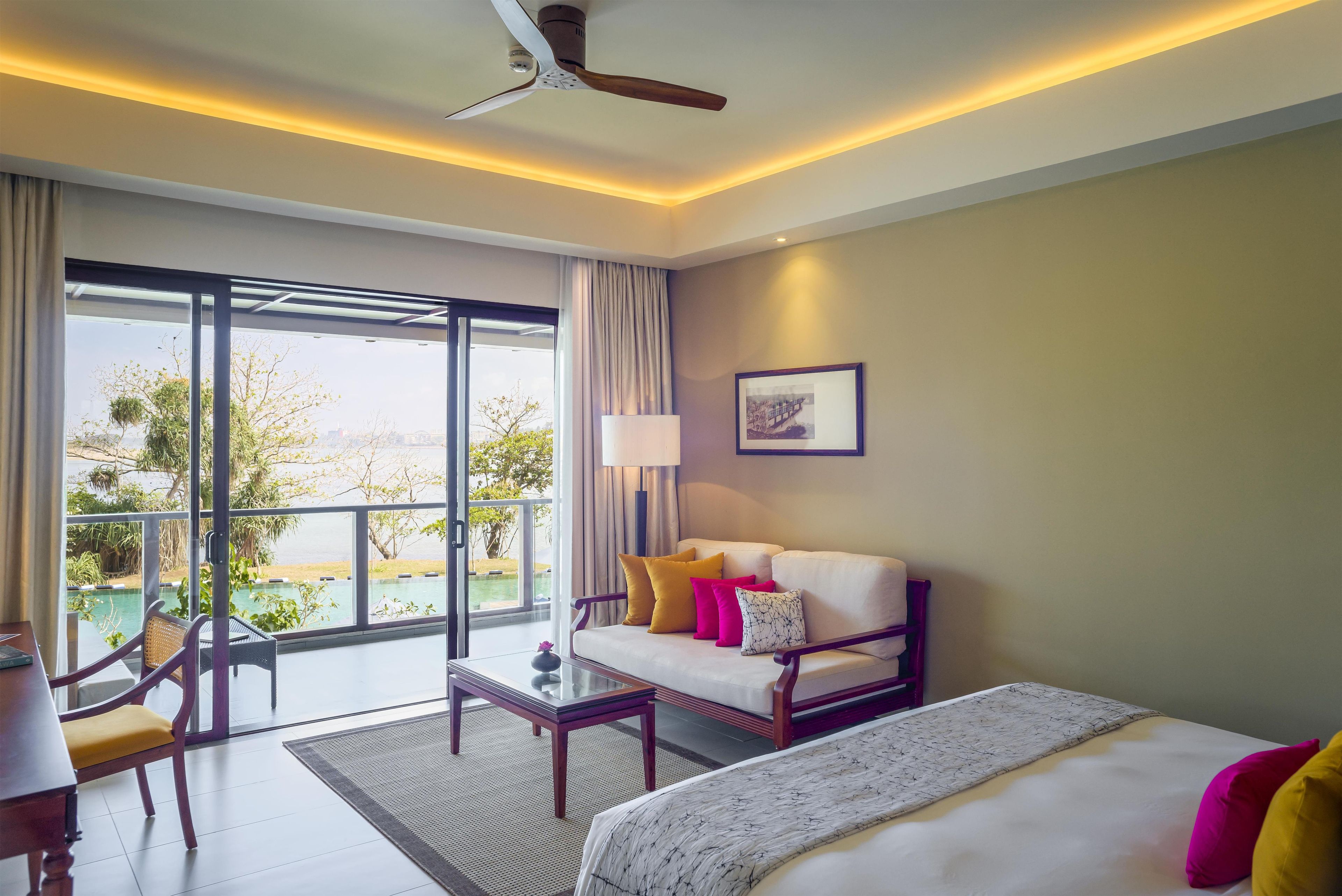 A hotel room with a bed, a sofa, a coffee table, a desk, and a view of a pool and greenery.