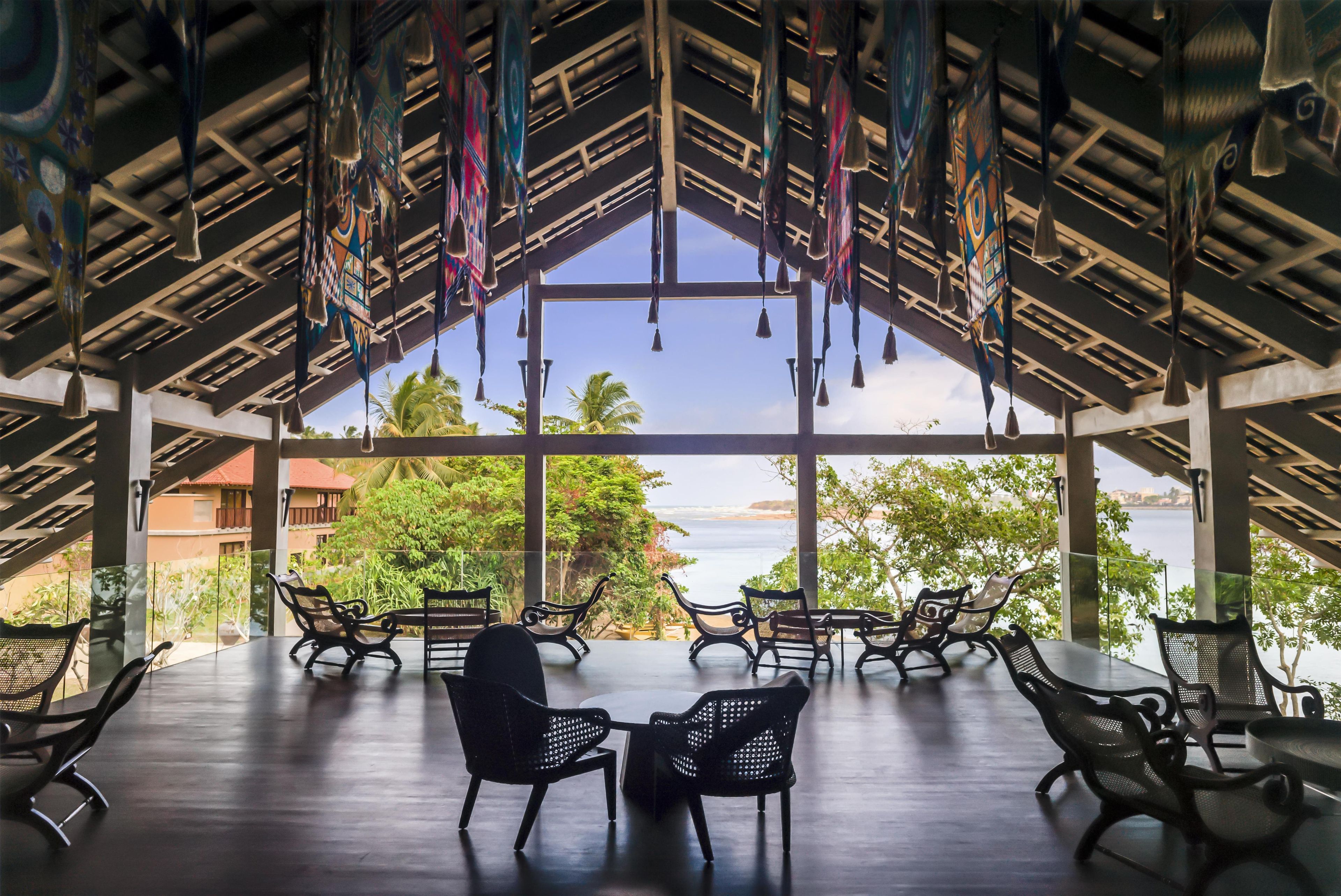 An open-air seating area with tables and chairs, a view of the sea, and a thatched roof structure.