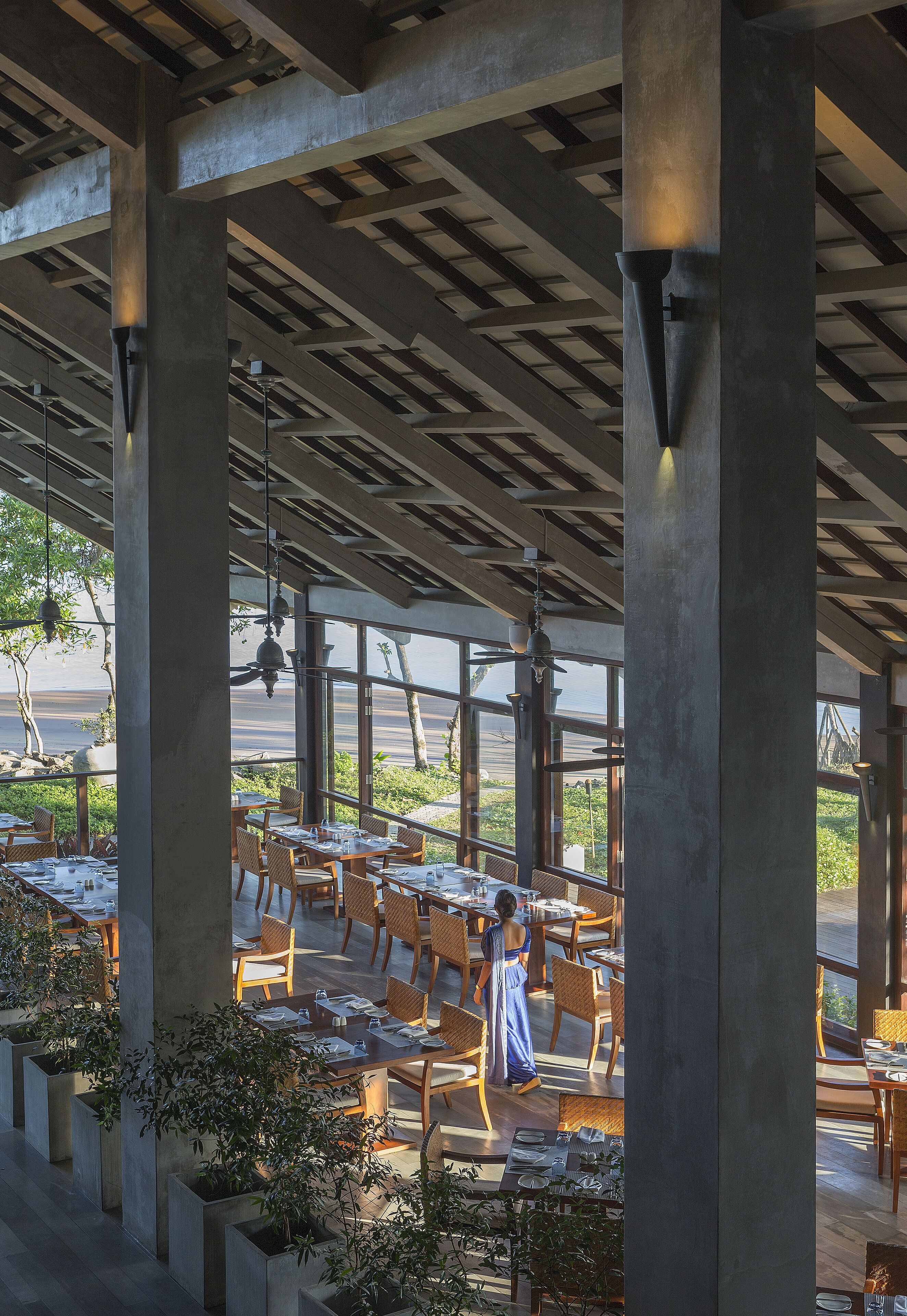 Un área de comedor al aire libre con mesas y sillas, grandes pilares de concreto y luces colgantes.