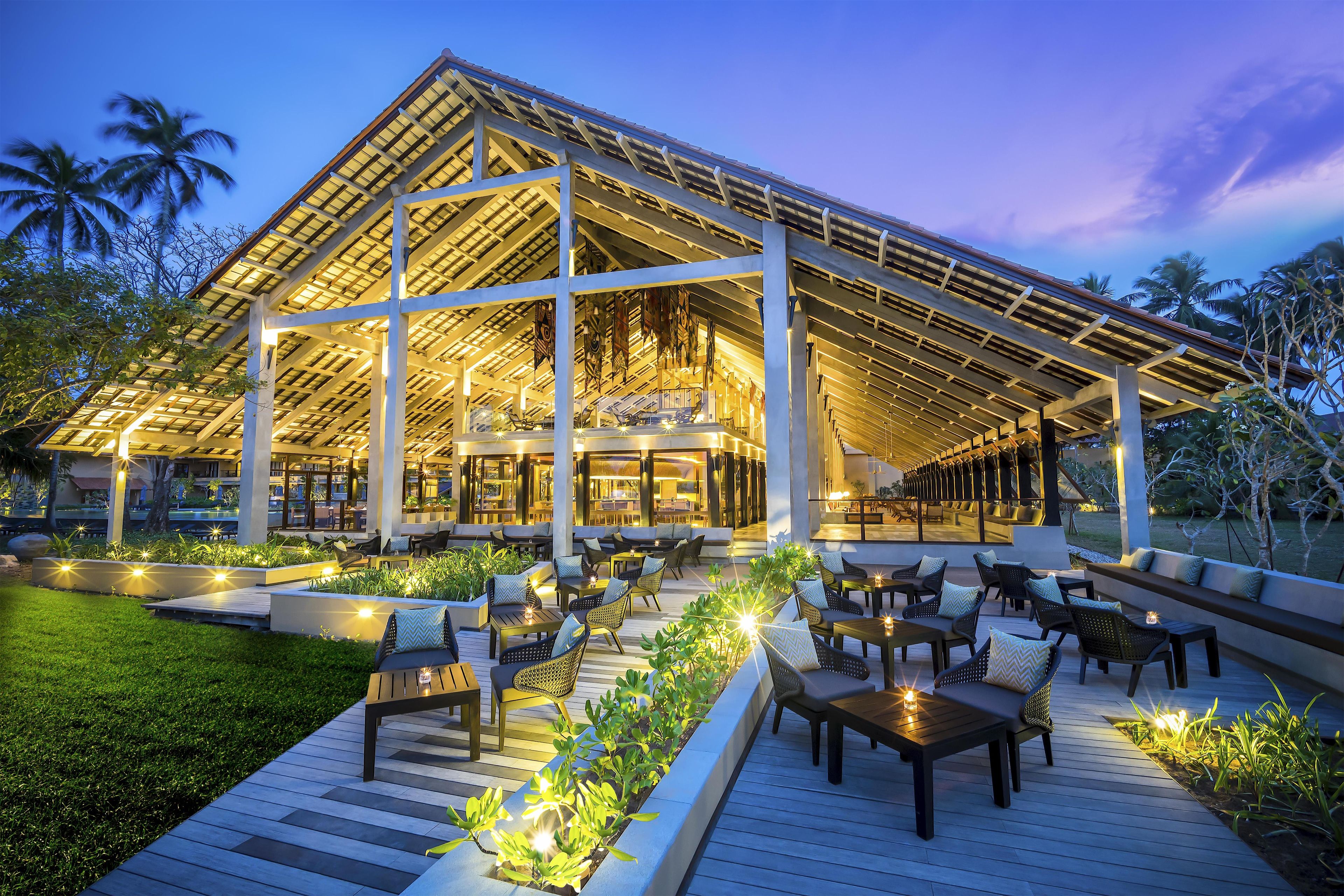 Een moderne, buitenloungeset met zitplaatsen en tafels, omgeven door groen en een overdekte constructie.