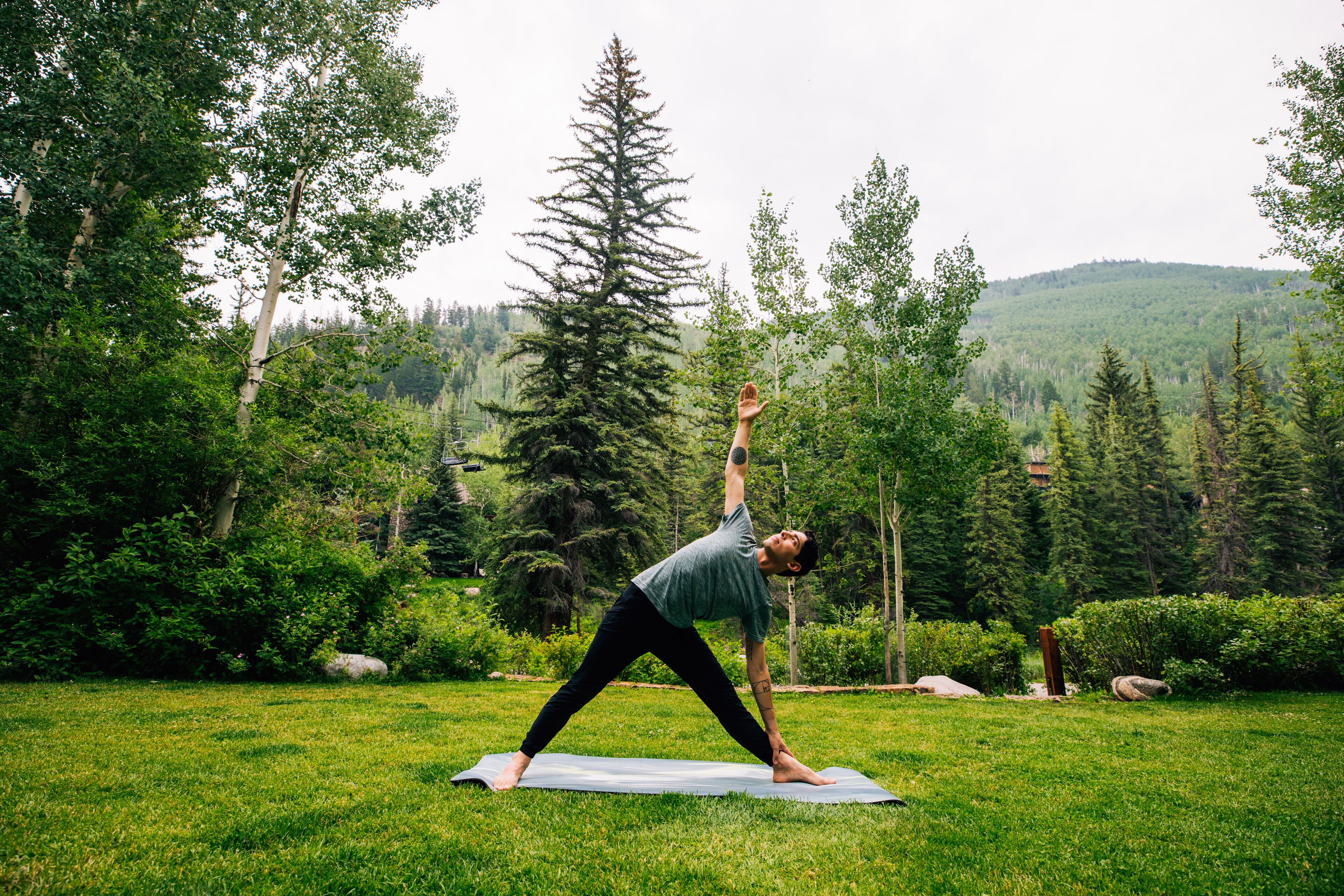 A person performing a yoga pose on a mat in a grassy outdoor area with trees and hills in the background.