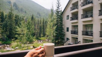 A hand holding a mug on a balcony overlooking a mountainous landscape with a building in the background.
