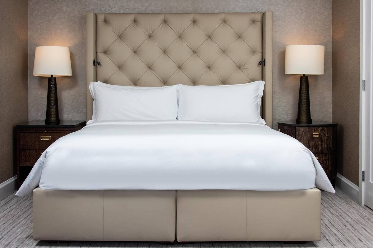 Ein großes Bett mit einem karrierten Kopfteil, zwei Nachttische mit Lampen und ein Teppich in neutralen Farben.