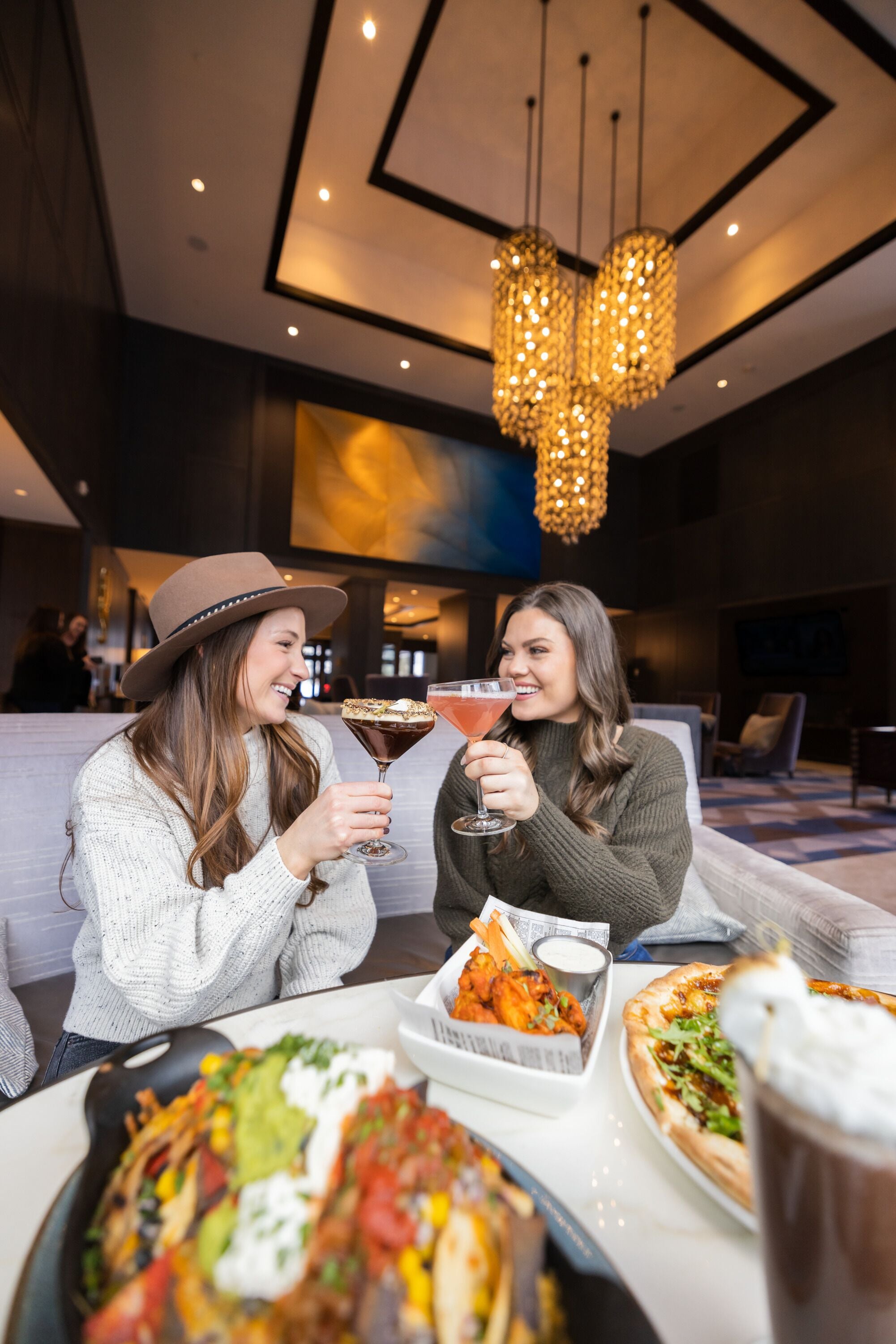 Two people sitting at a table with a variety of food and drinks, including nachos, a pizza, and a martini.