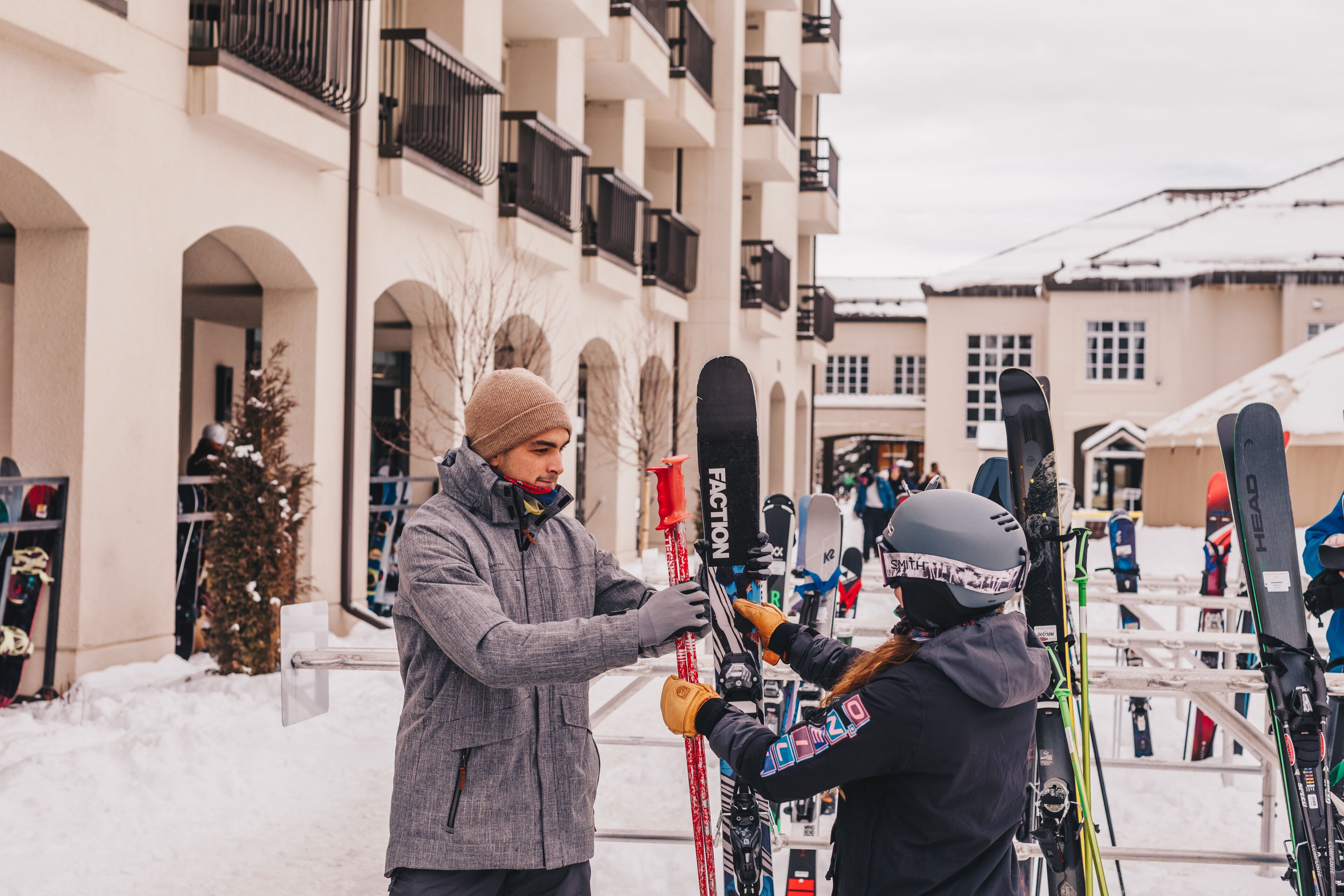 A person adjusting skis at a ski rental area with a building in the background.