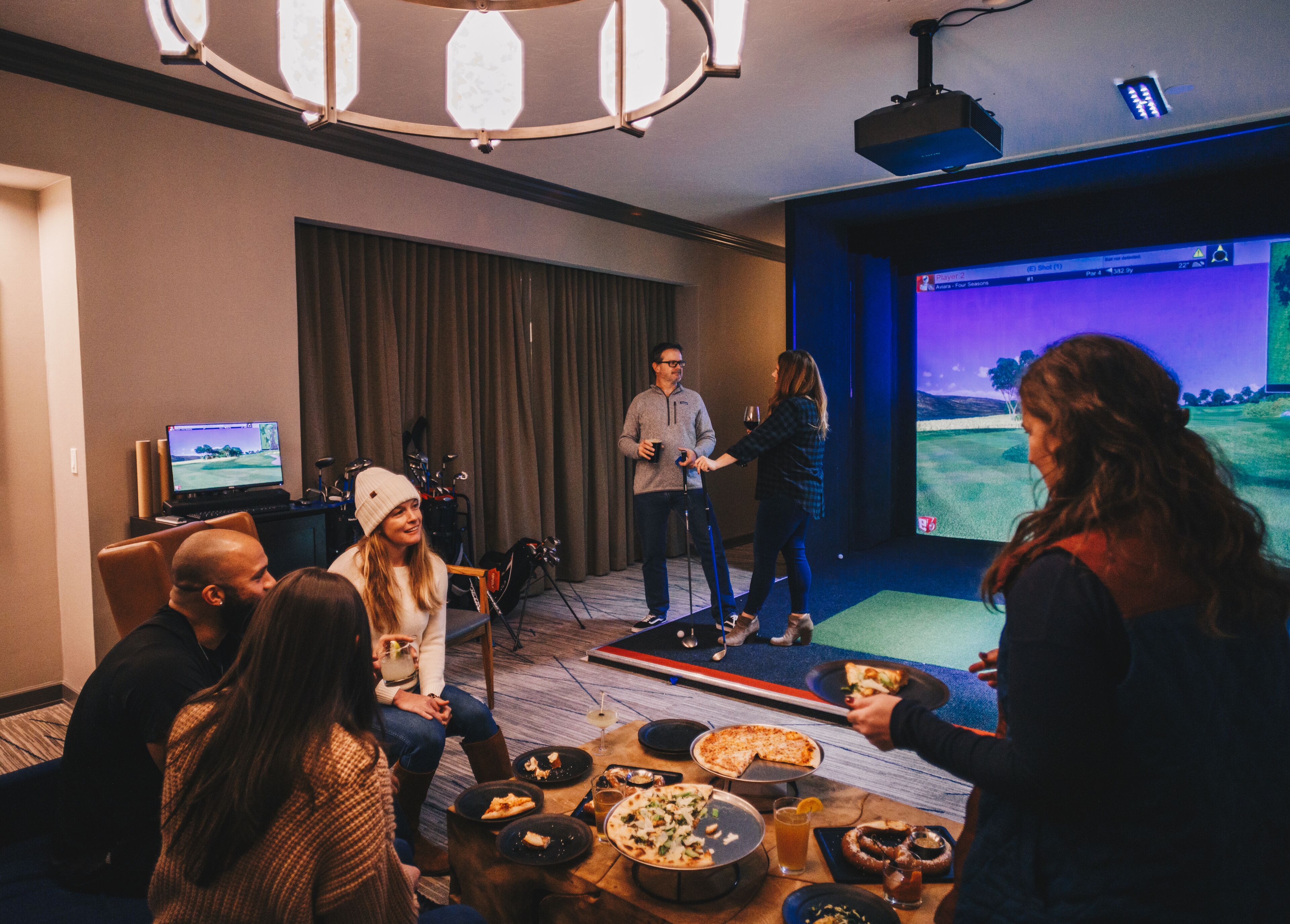 En gruppe mennesker i et rum med et stort skærmbillede af en golfkamp, et bord med mad og drikkevarer, og et afslappet siddeområde.