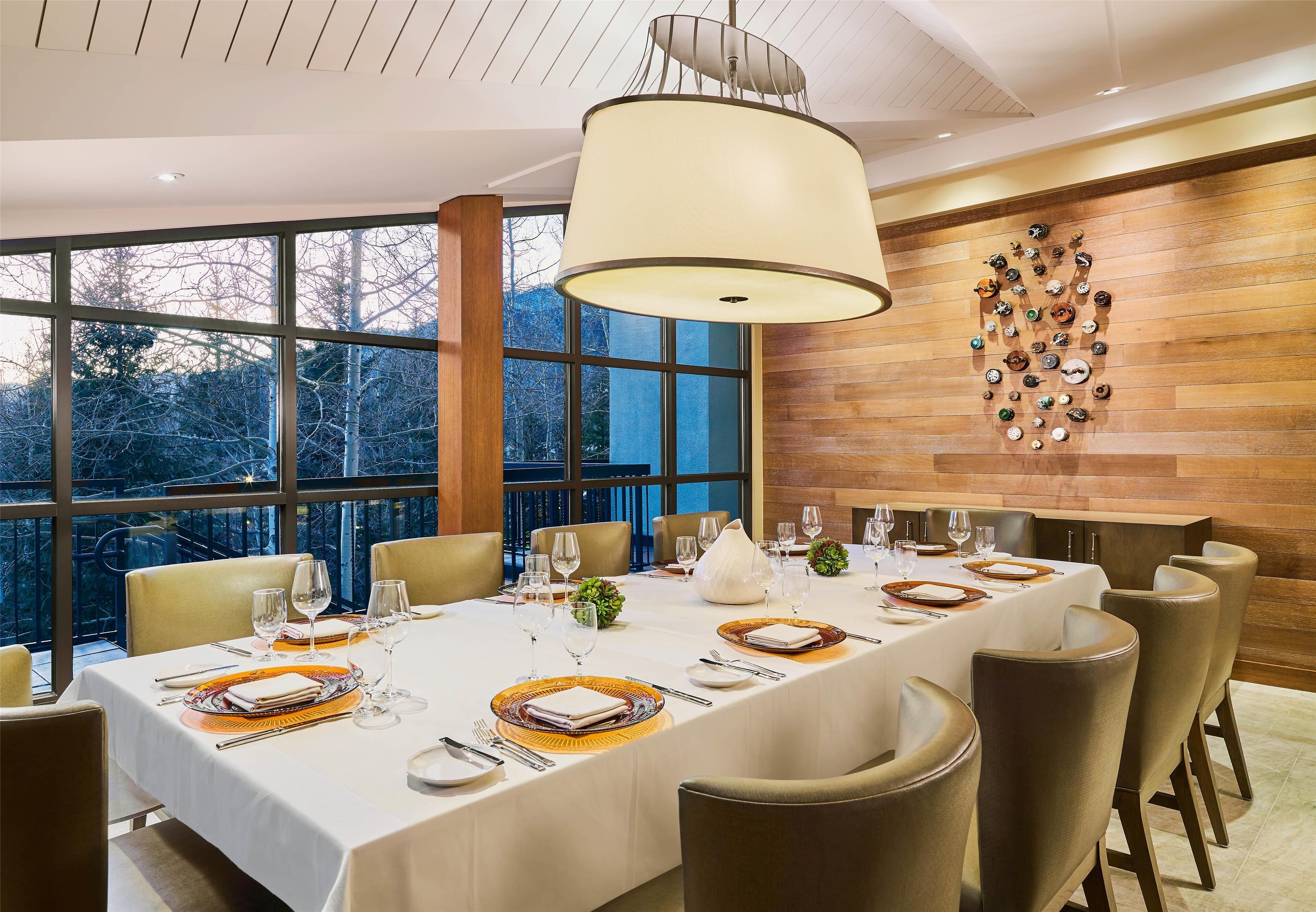 A dining area with a long table set for a meal, large windows, and a wooden wall with a decorative plate arrangement.