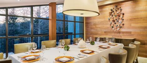 A dining area with a long table set for a meal, large windows, and a wooden wall with a decorative plate arrangement.
