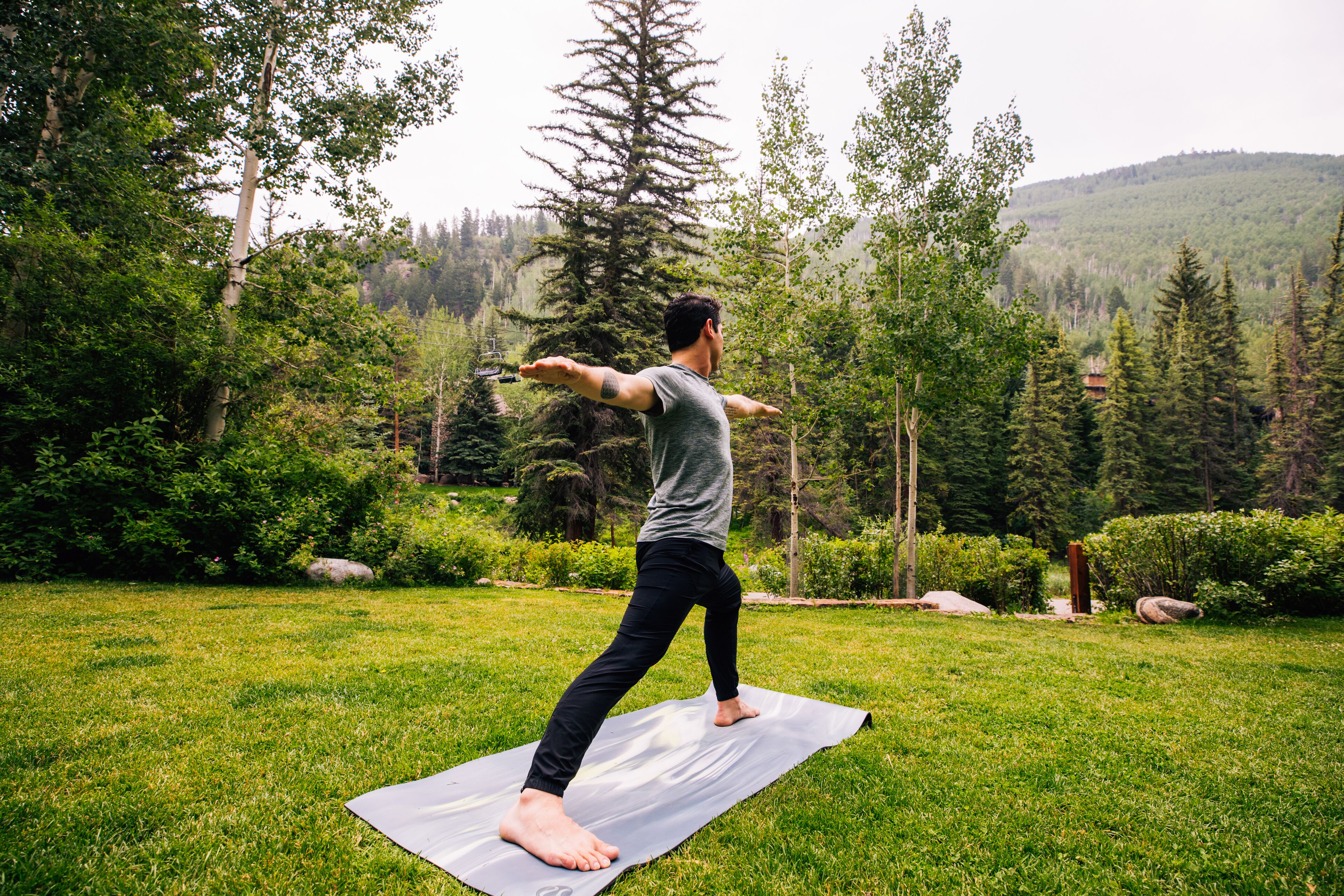 Una persona practicando yoga sobre una colchoneta en un área con césped, con árboles y colinas al fondo.