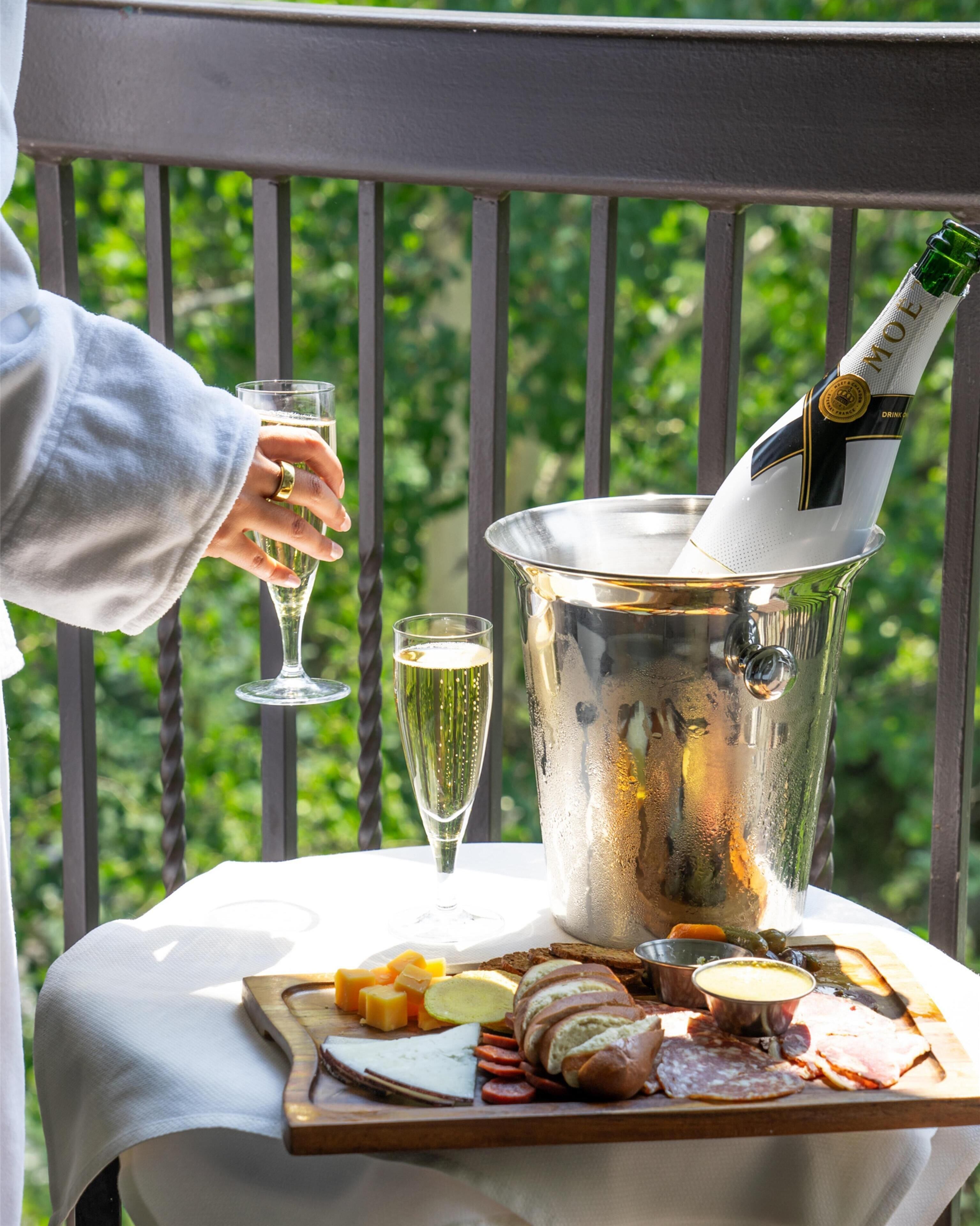 A person holding a champagne glass, a bottle in an ice bucket, and a charcuterie board on a balcony.