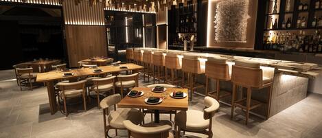 A modern bar area with wooden tables and chairs, a bar counter, and a wall of liquor bottles.