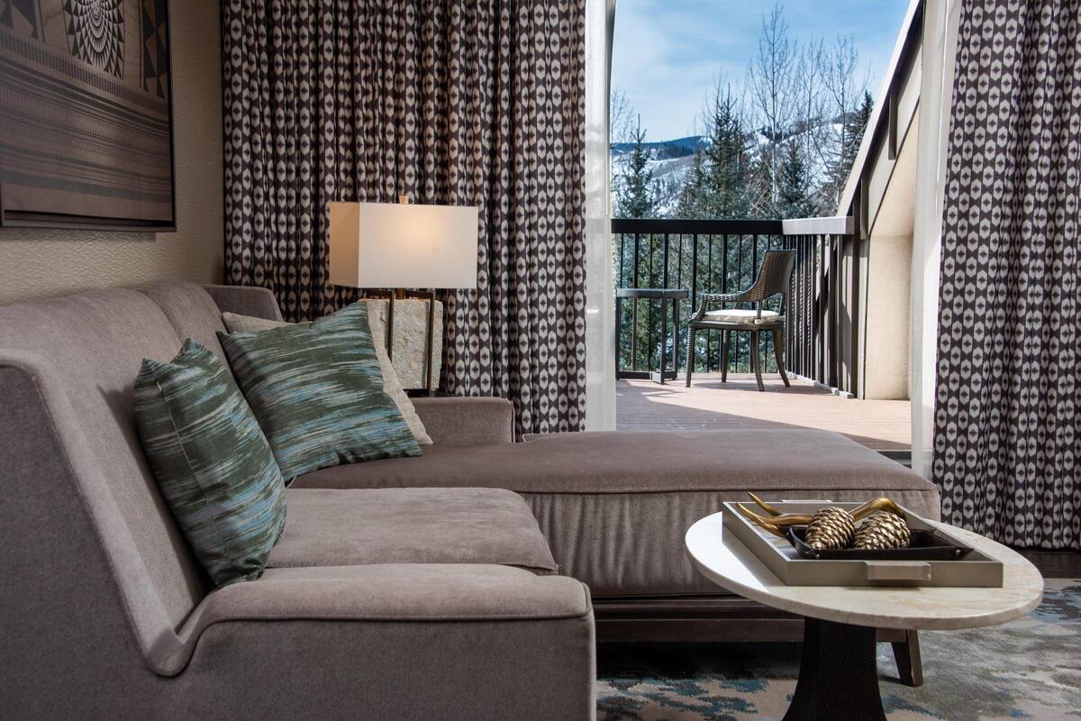 Ein modernes Wohnzimmer mit einer Couch, einem Beistelltisch mit Dekoration, gemusterten Vorhängen und Blick auf einen Balkon mit Gartenmöbeln.