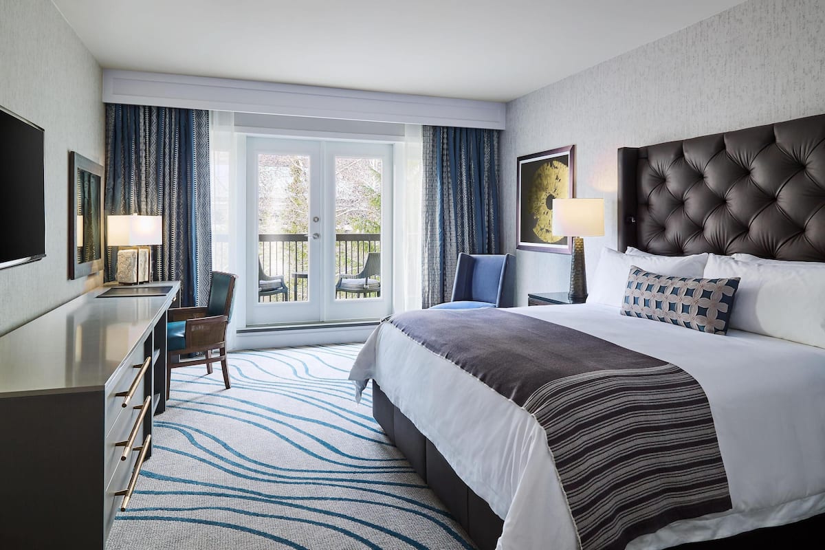 Ein Hotelzimmer mit einem großen Bett, einem Schreibtisch, einem Sessel, einem Fernseher und einem Balkon mit Vorhängen.