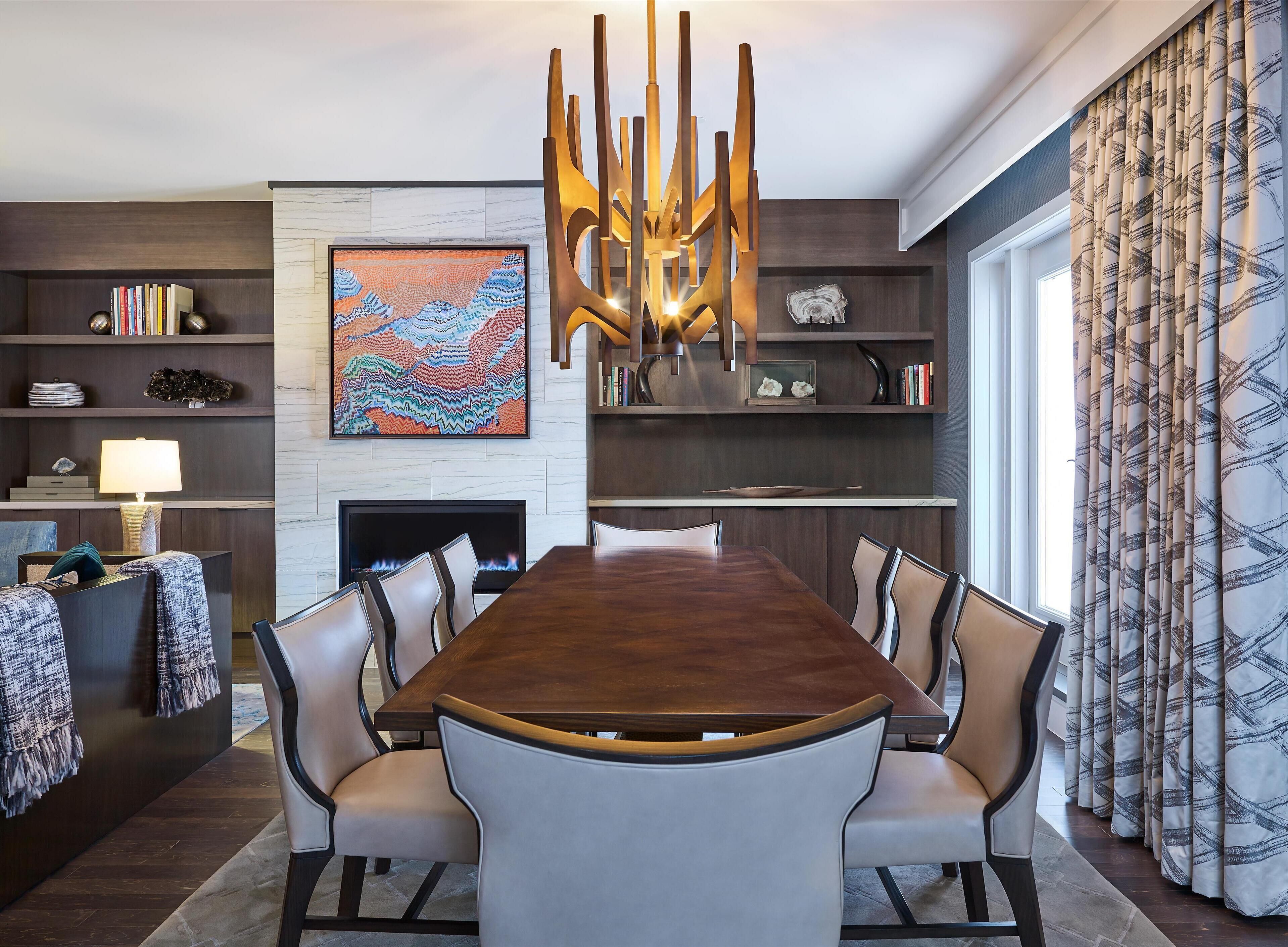 A modern dining area with a large wooden table, cushioned chairs, a fireplace, bookshelves, and a decorative wall piece.