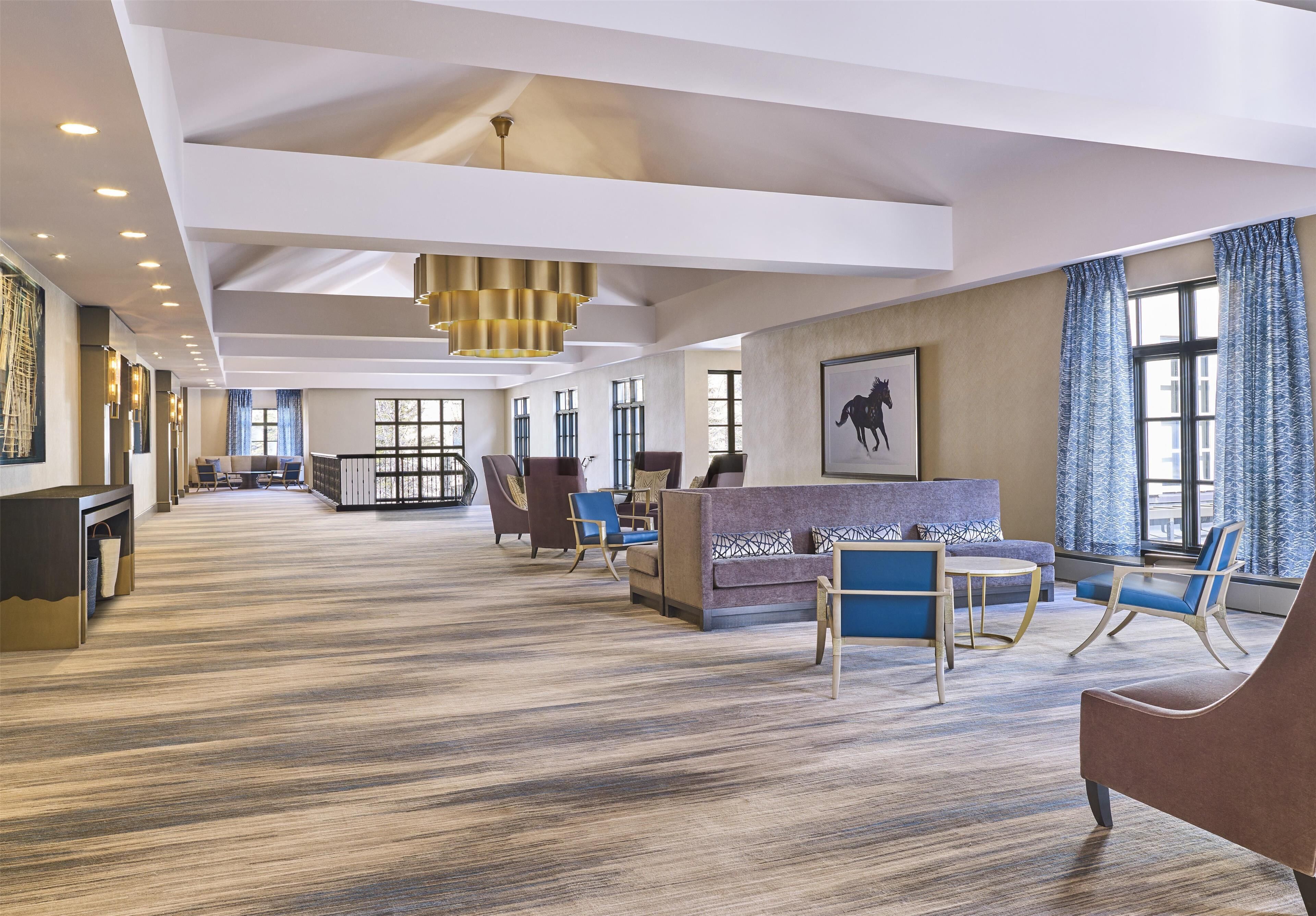 Un vestíbulo amplio con alfombra estampada, una recepción, zona de estar con sillas y sofás, ventanas grandes con cortinas e iluminación empotrada en el techo.