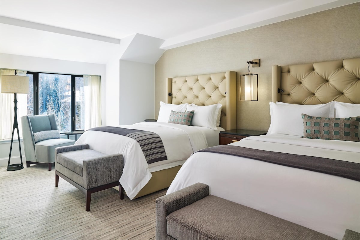 Ein Hotelzimmer mit zwei Betten, einer Sitzecke mit einem Sessel, einer Lampe und Blick auf Bäume durch das Fenster.