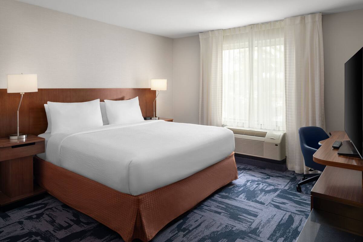 A hotel room with a large bed, two bedside tables with lamps, a desk with a chair, and a large window with curtains.