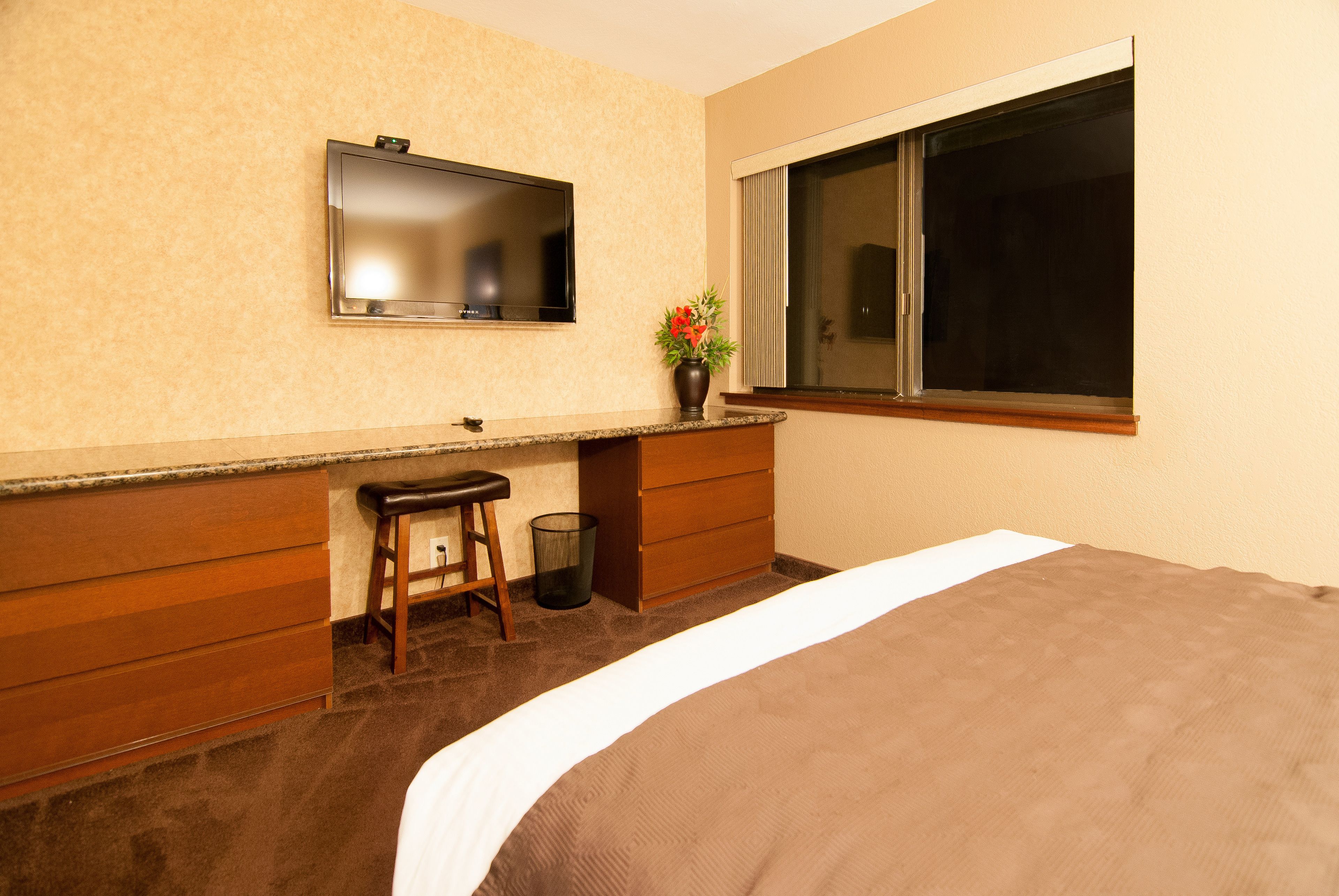 A hotel room with a bed, a wooden dresser, a vanity with a mirror, a small stool, a trash can, and a window with blinds.