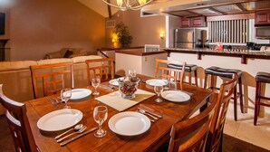 Zona pranzo con tavolo in legno apparecchiato per sei persone, circondato da sedie. Sullo sfondo si intravede una cucina con elettrodomestici moderni.