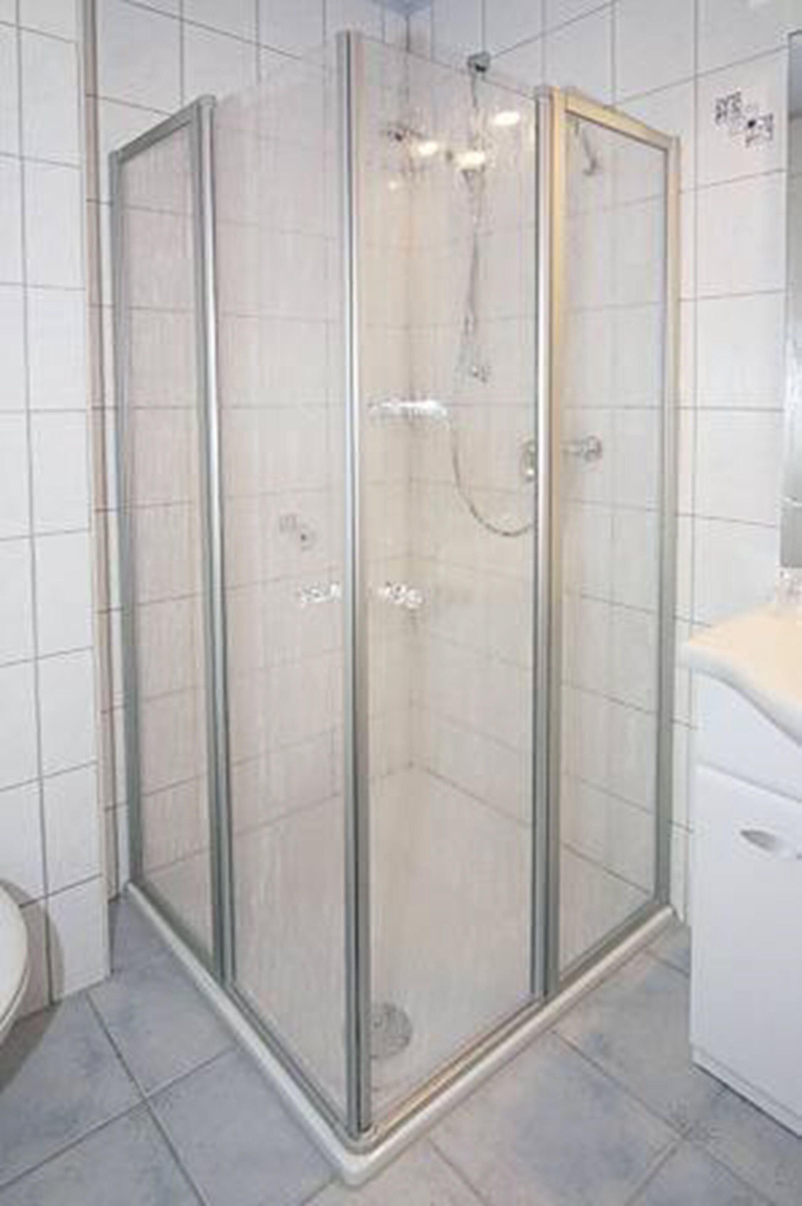A glass-enclosed shower stall with a white tiled floor and a white sink to the right.
