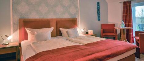 A double bed with a wooden headboard, a red throw blanket, two bedside tables with lamps, a red armchair, and a small table with a bottle and glasses.