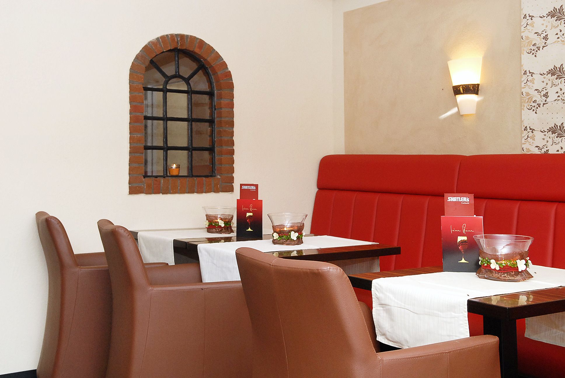 A restaurant interior with red booths, a brick arched window, and tables with menus and candles.